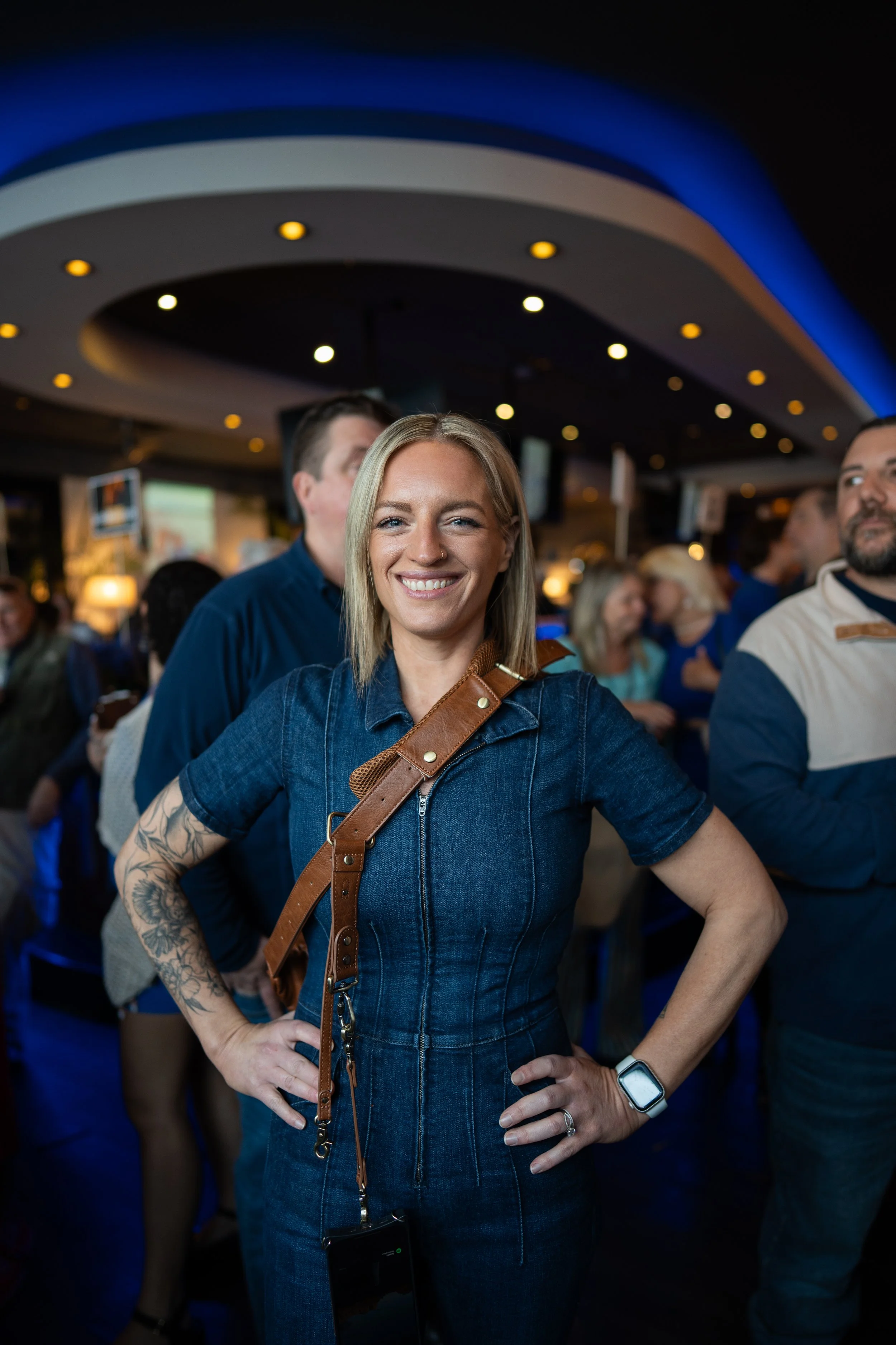 A smiling blonde woman wearing a denim jumpsuit and a smartwatch, standing with her hands on her hips in a crowded indoor setting with people in the background.