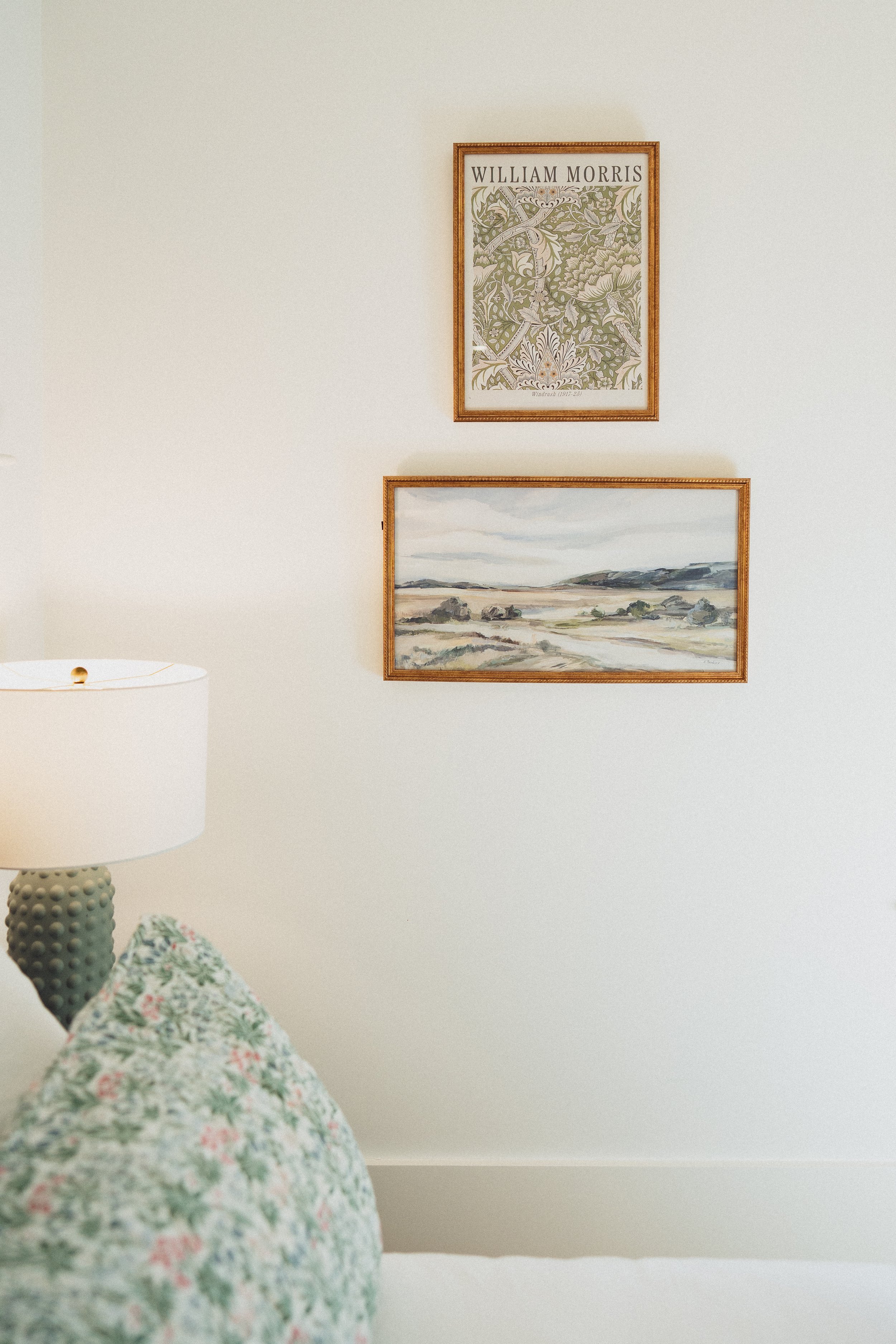 Interior of a room showing two framed pictures on the wall, a partially visible bed with a floral pillow, and a table lamp with a textured green base.
