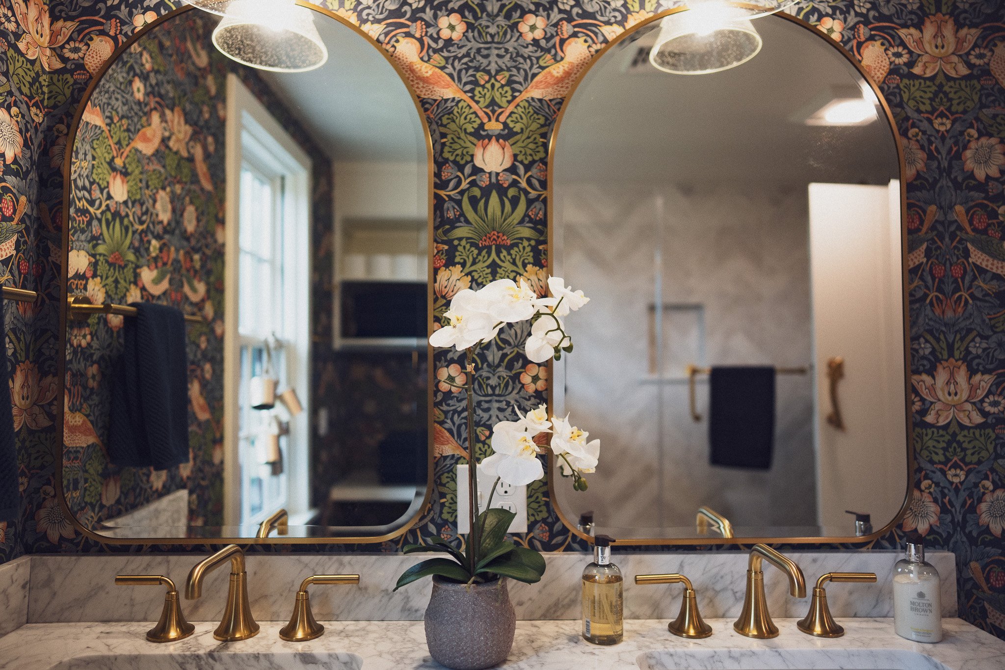 A bathroom with a marble countertop, two brass faucets, and a potted white orchid flower. There are two round mirrors with gold frames and floral-patterned wallpaper in dark colors. A soap dispenser and hand lotion are on the counter, and a window wi