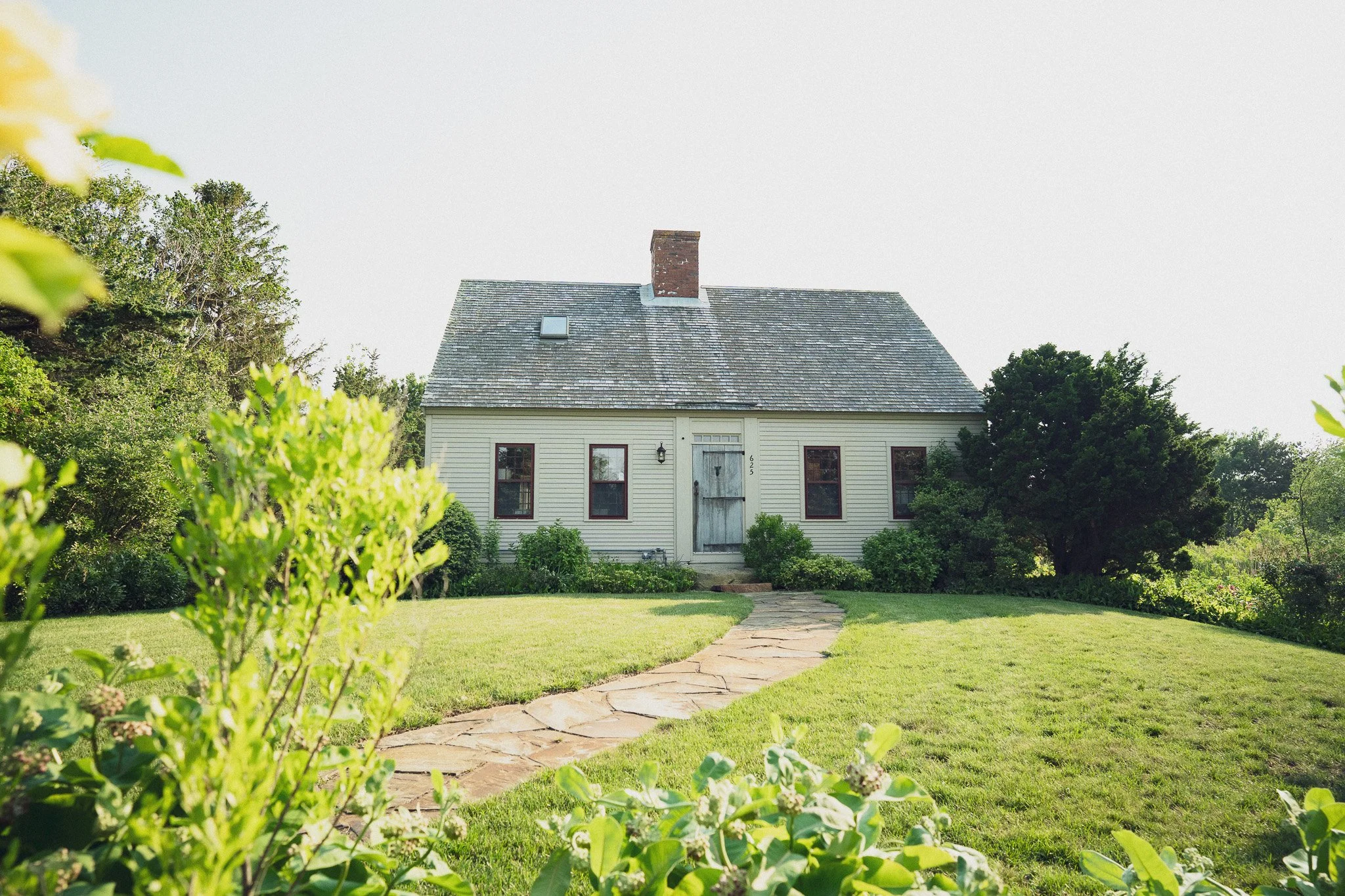 A white house with a shingled roof, four windows, a chimney, and a wooden door, surrounded by green bushes, a lawn, and trees, with a stone pathway leading to the door.