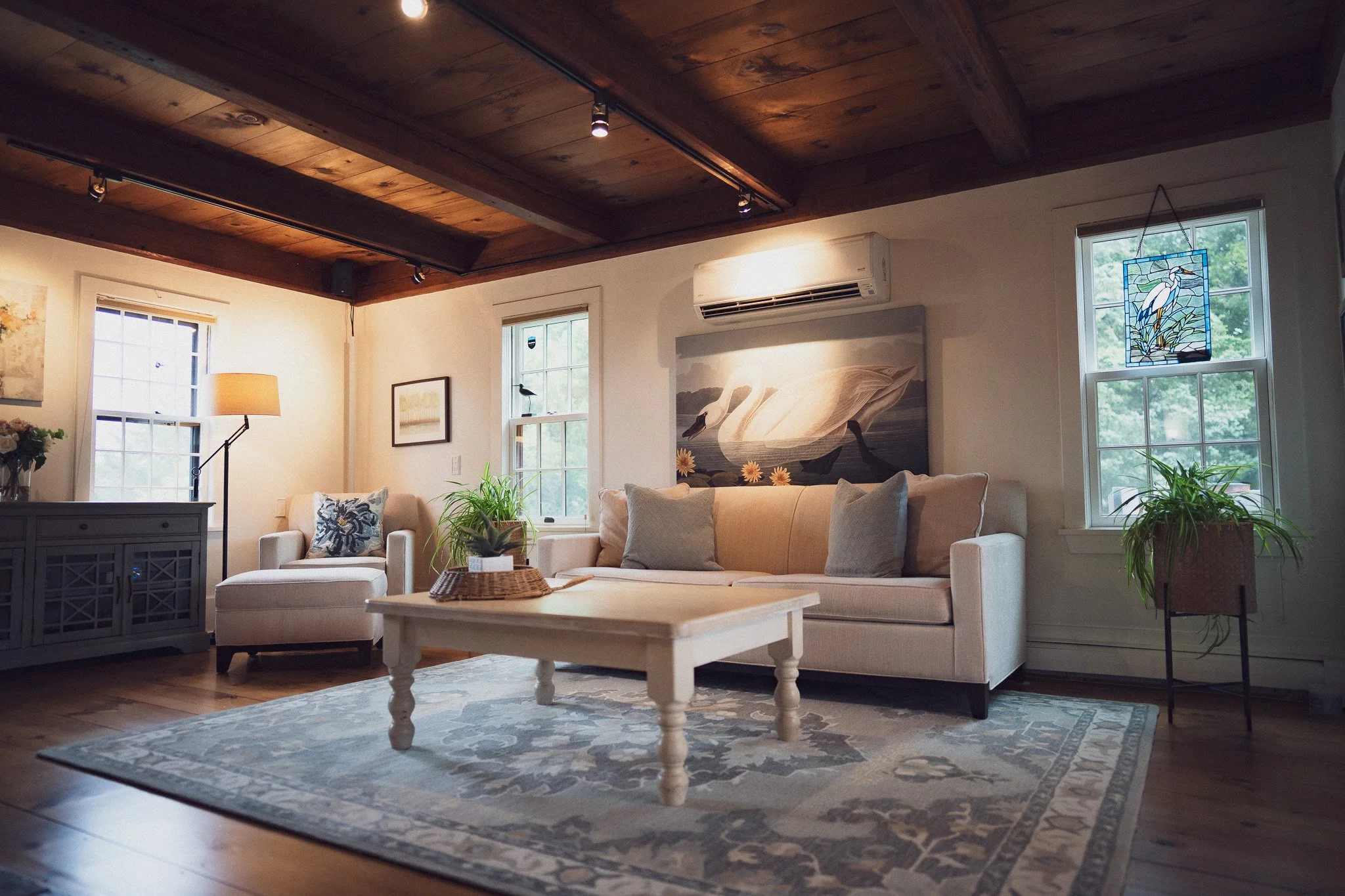 Living room with white couch, armchair, coffee table, and houseplants, featuring wooden ceiling beams and large windows with white frames.