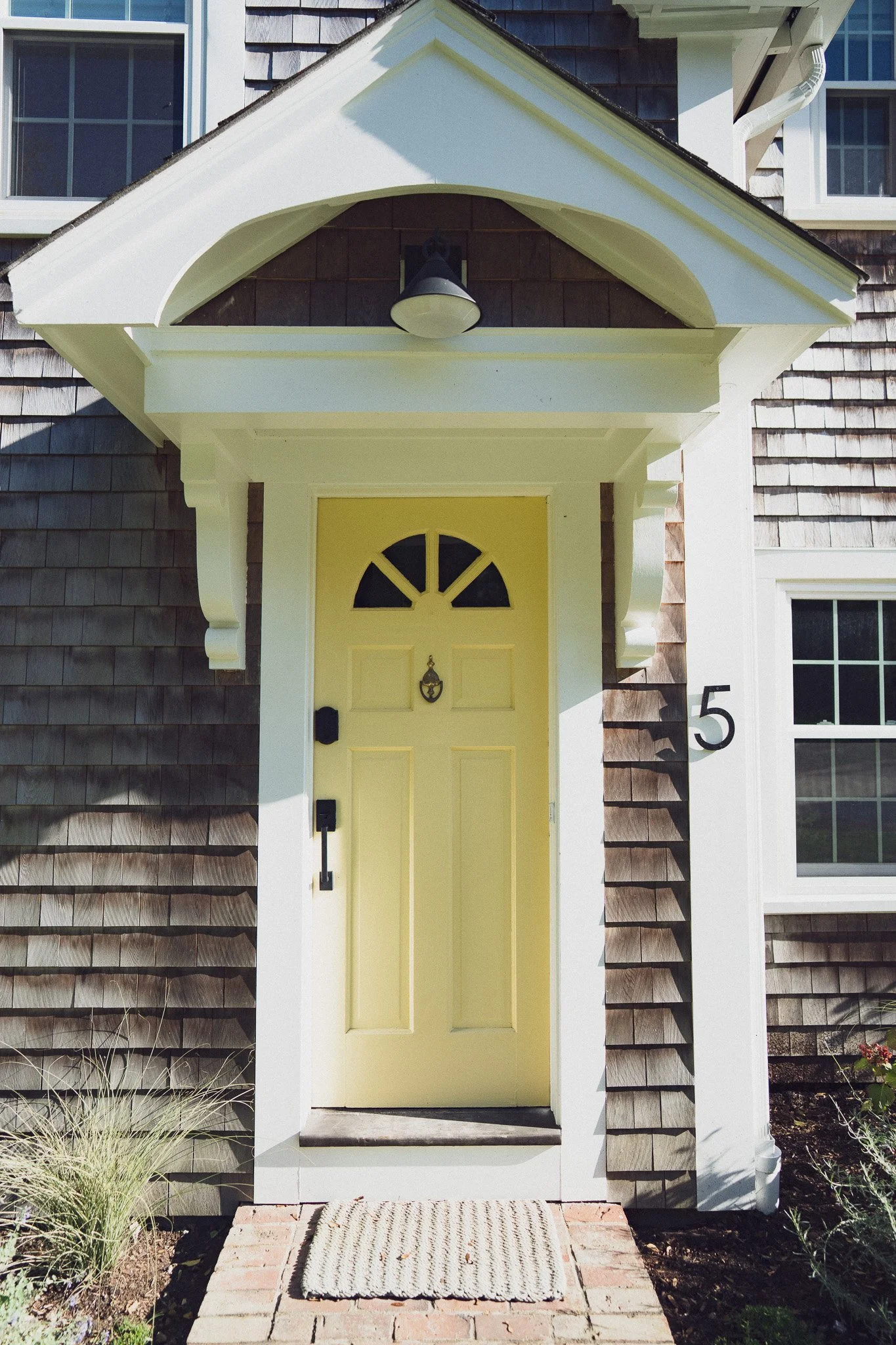 Close-up of a yellow front door with a half-moon window, black hardware, and a door knocker, surrounded by white trim and a small brick step with a striped doormat. Part of a house with shingled siding and white window frames is visible.