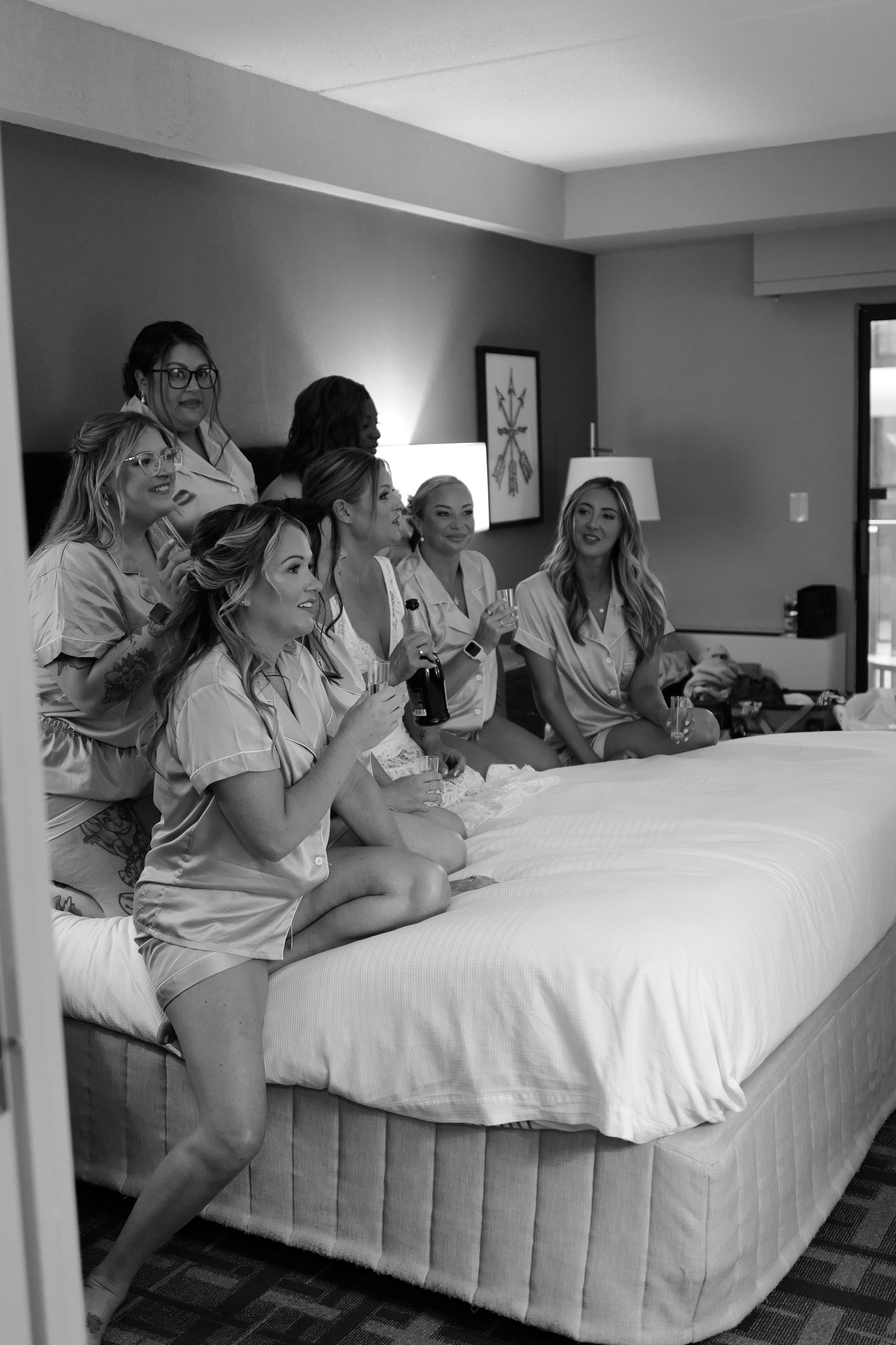 Group of women in pajamas sitting on a bed, celebrating with drinks in a hotel room.