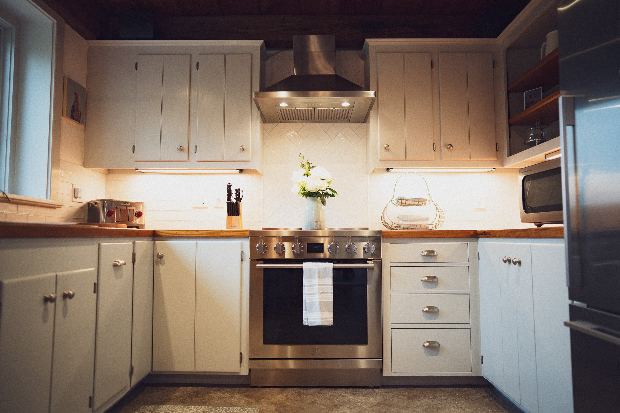 Kitchen with white cabinets, wooden countertops, stainless steel oven, range hood, and small appliances, including a toaster and microwave, with a vase of white flowers on the counter.