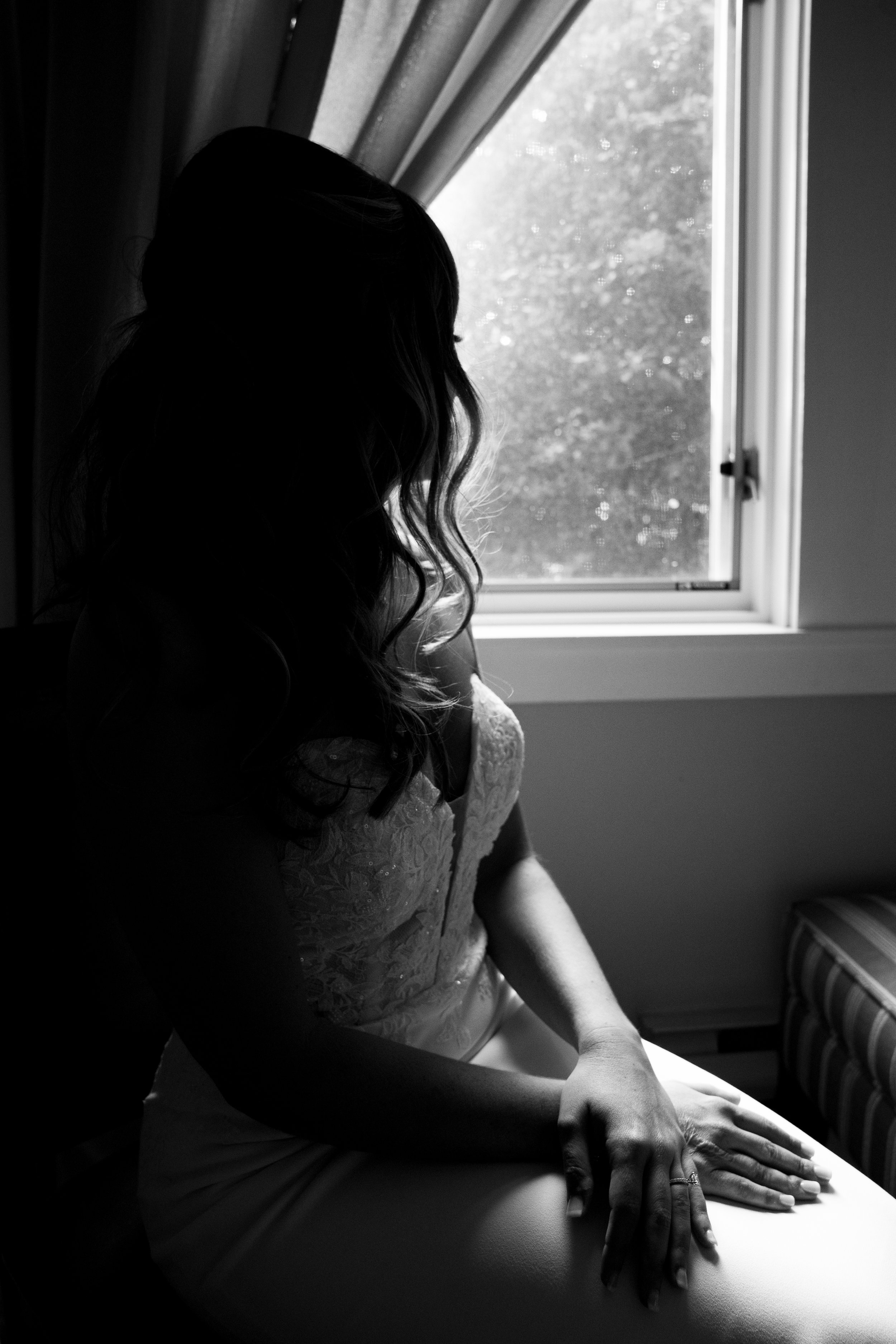 A woman with long, wavy hair sitting by a window, dressed in a lace top and skirt, looking outside, in black and white.