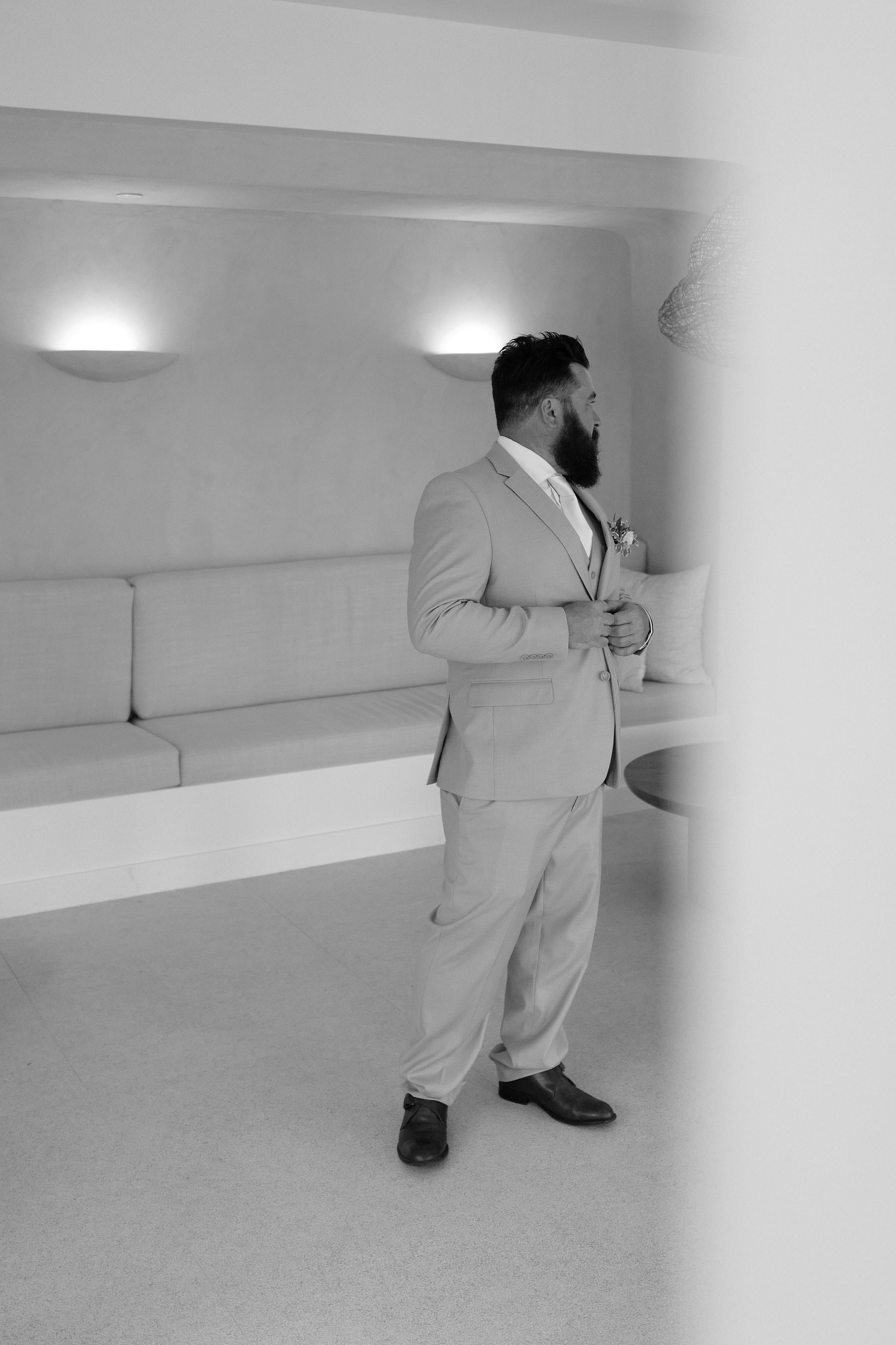 A man with a beard in a light-colored suit standing in a room, looking to the right, with a bench with cushions and wall lights in the background.