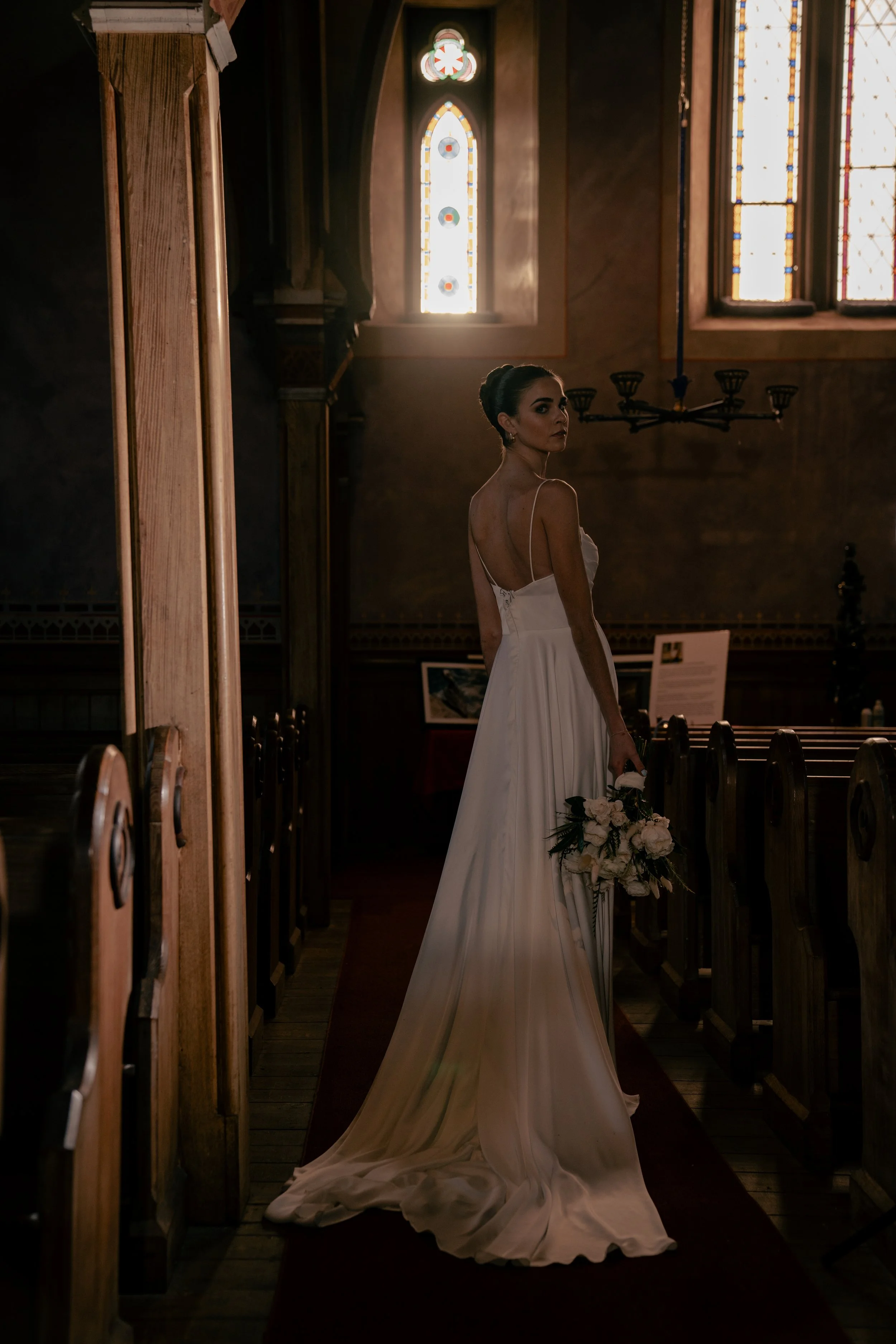 A bride in a white wedding dress holding a bouquet, standing inside a church with stained glass windows.