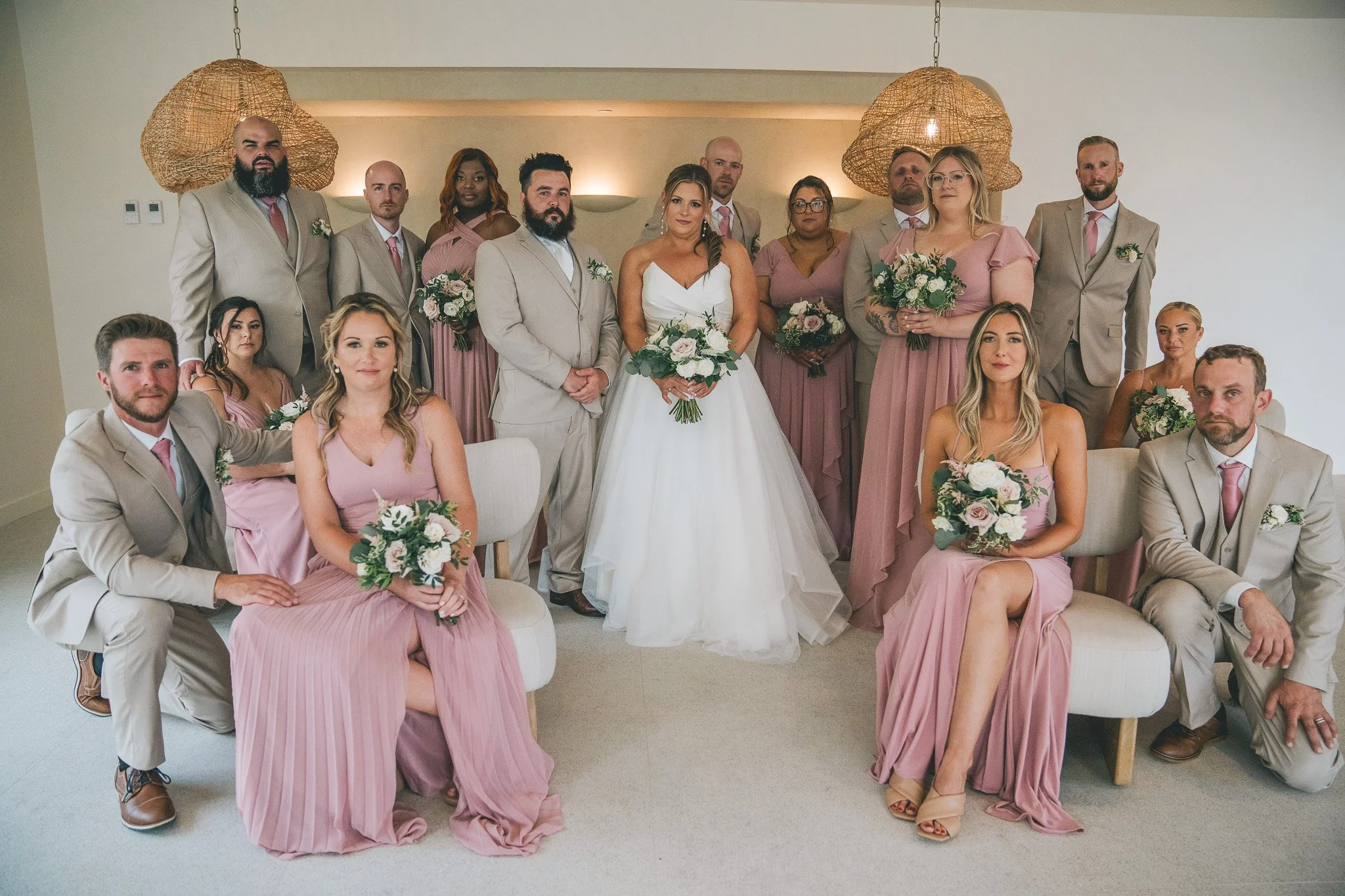 Group of wedding party members, including bride in white gown and bridesmaids in pink dresses, posing indoors with bouquets, with beige suits and a neutral background.