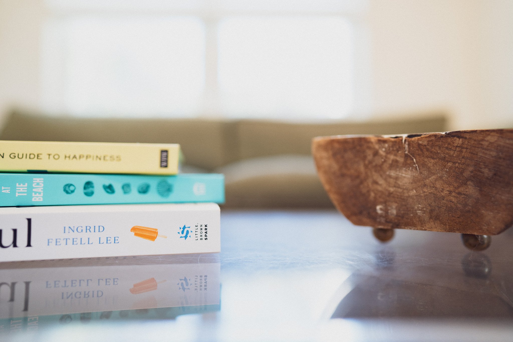 Stack of three colorful books with titles partially visible, a rustic wooden bowl on short legs, and a highly reflective surface creating a mirror image of the books.