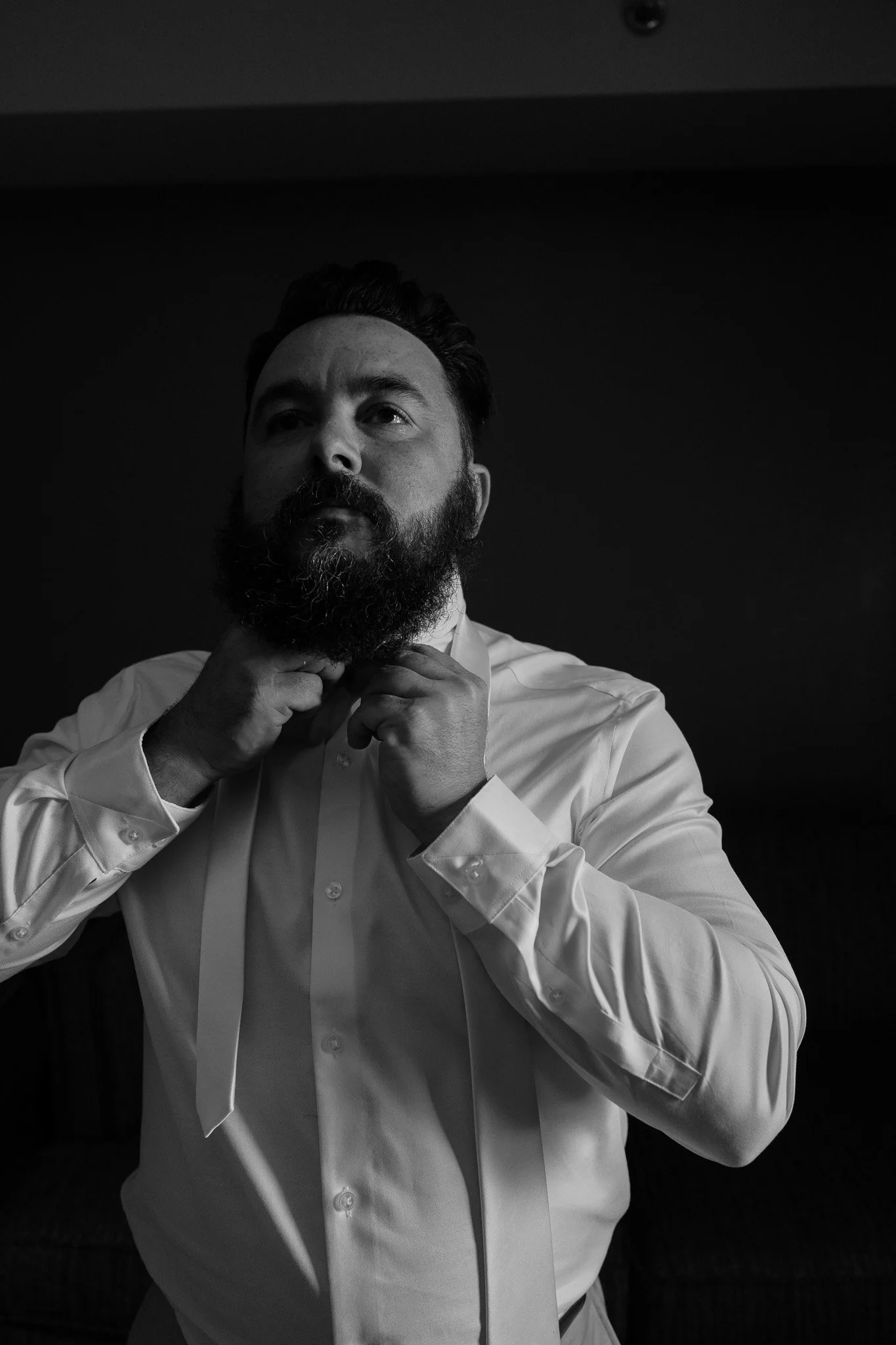 A man with a beard and short hair buttons his shirt collar in front of a dark background.