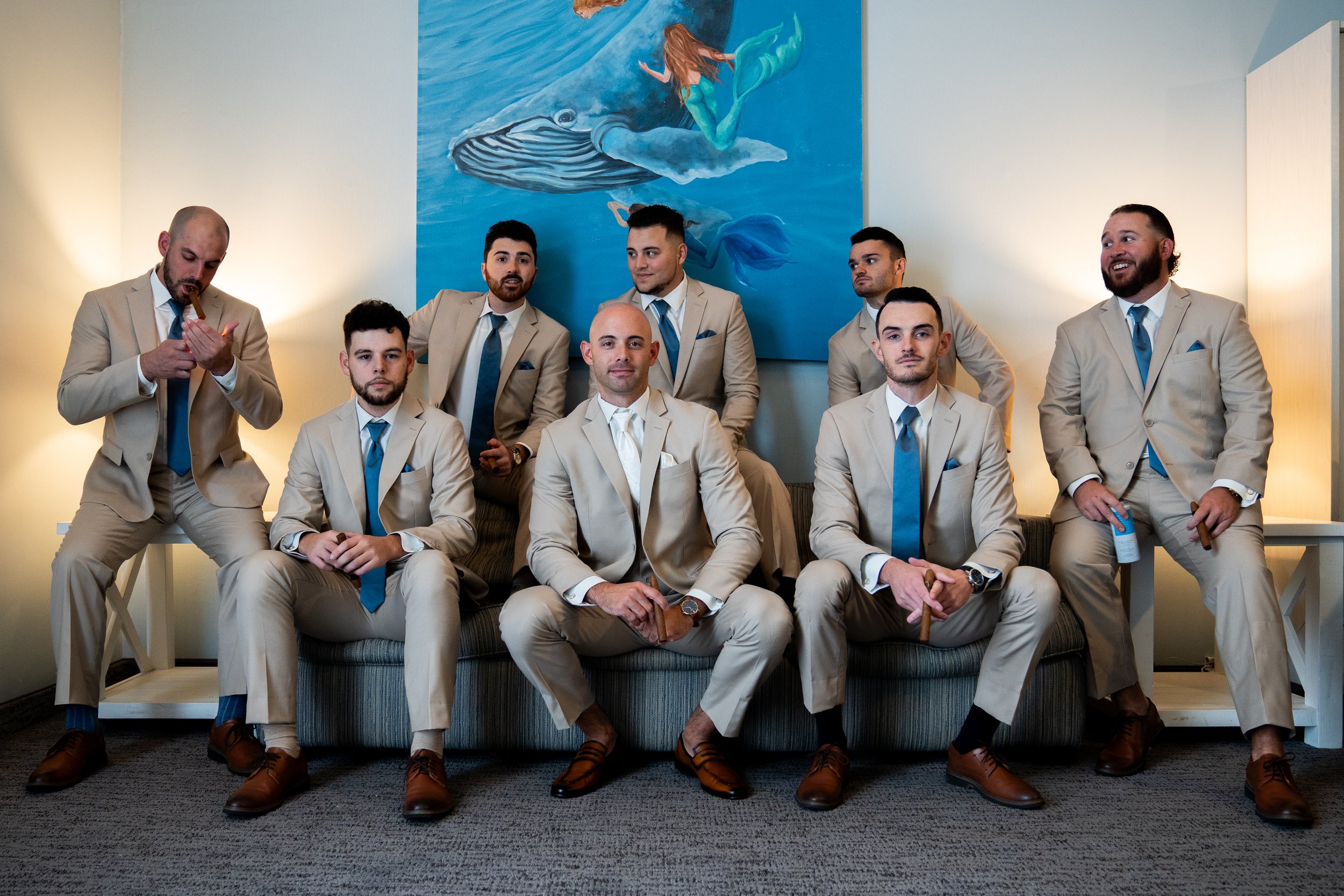 A group of nine men dressed in matching beige suits and blue ties, posed together in a room with a whale painting on the wall behind them.