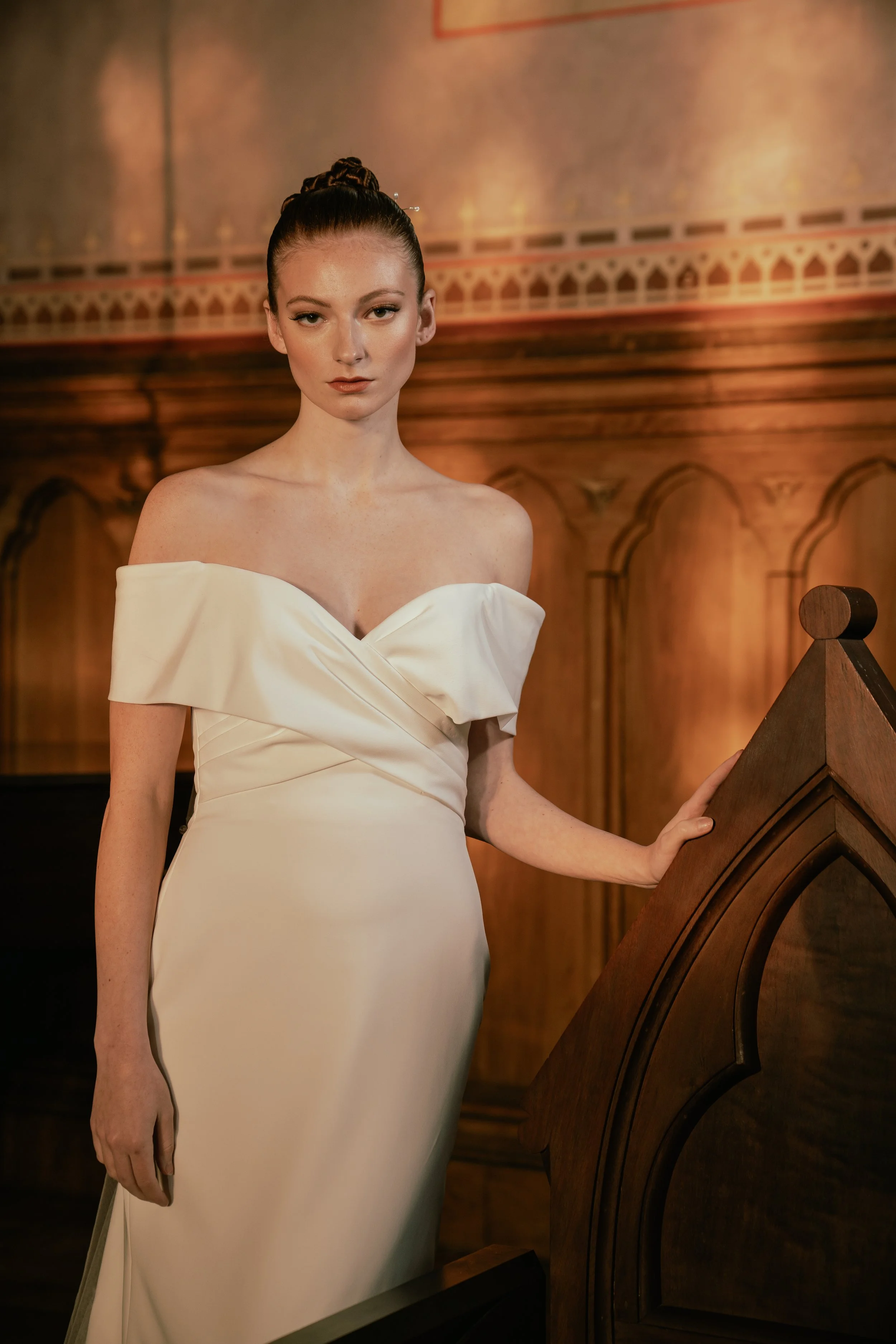 A woman in a white off-the-shoulder gown standing in a wood-paneled room.