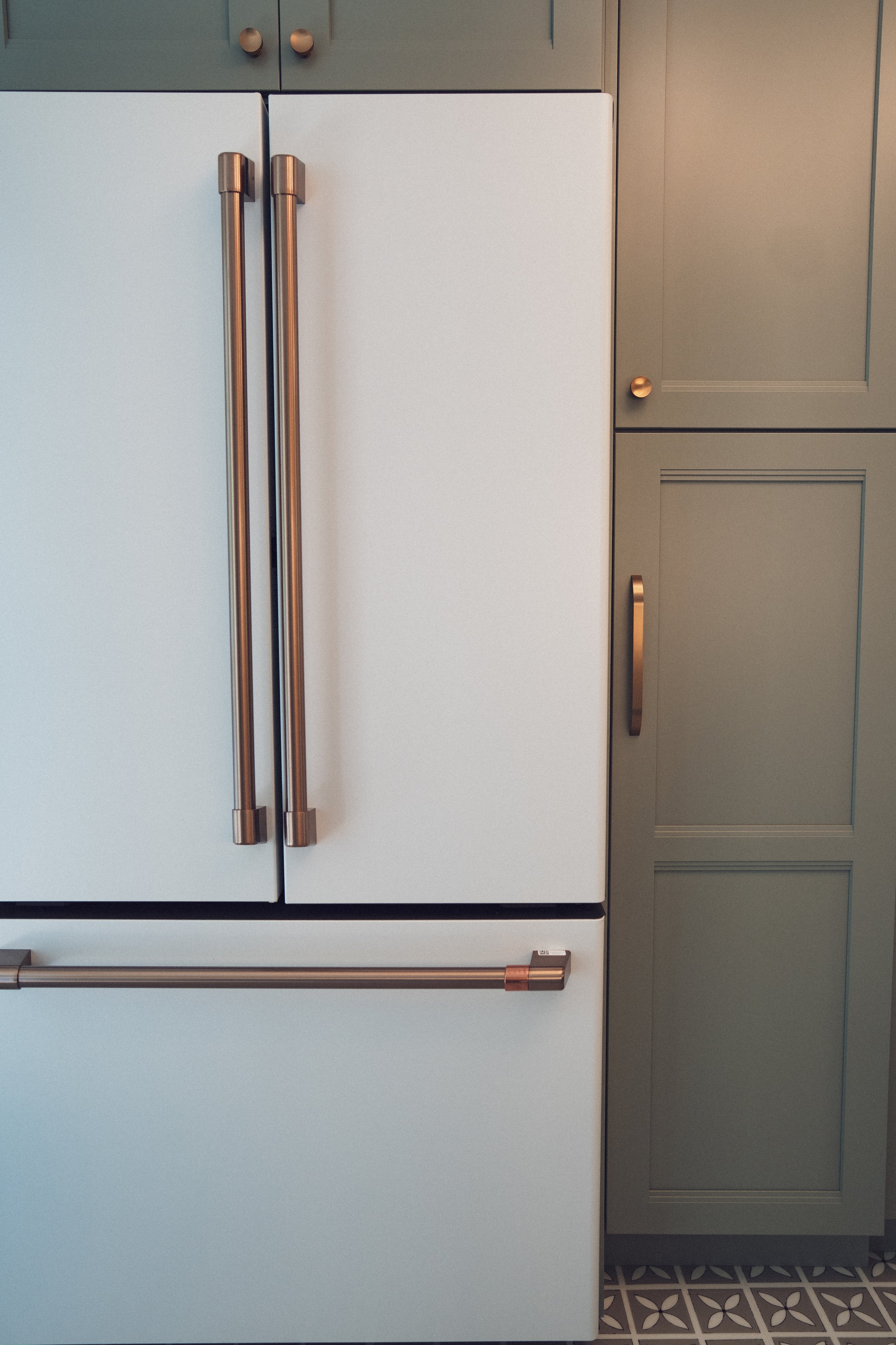 Close-up of a white refrigerator with stainless steel handles in a kitchen next to green cabinetry with gold knobs, and patterned floor tiles.