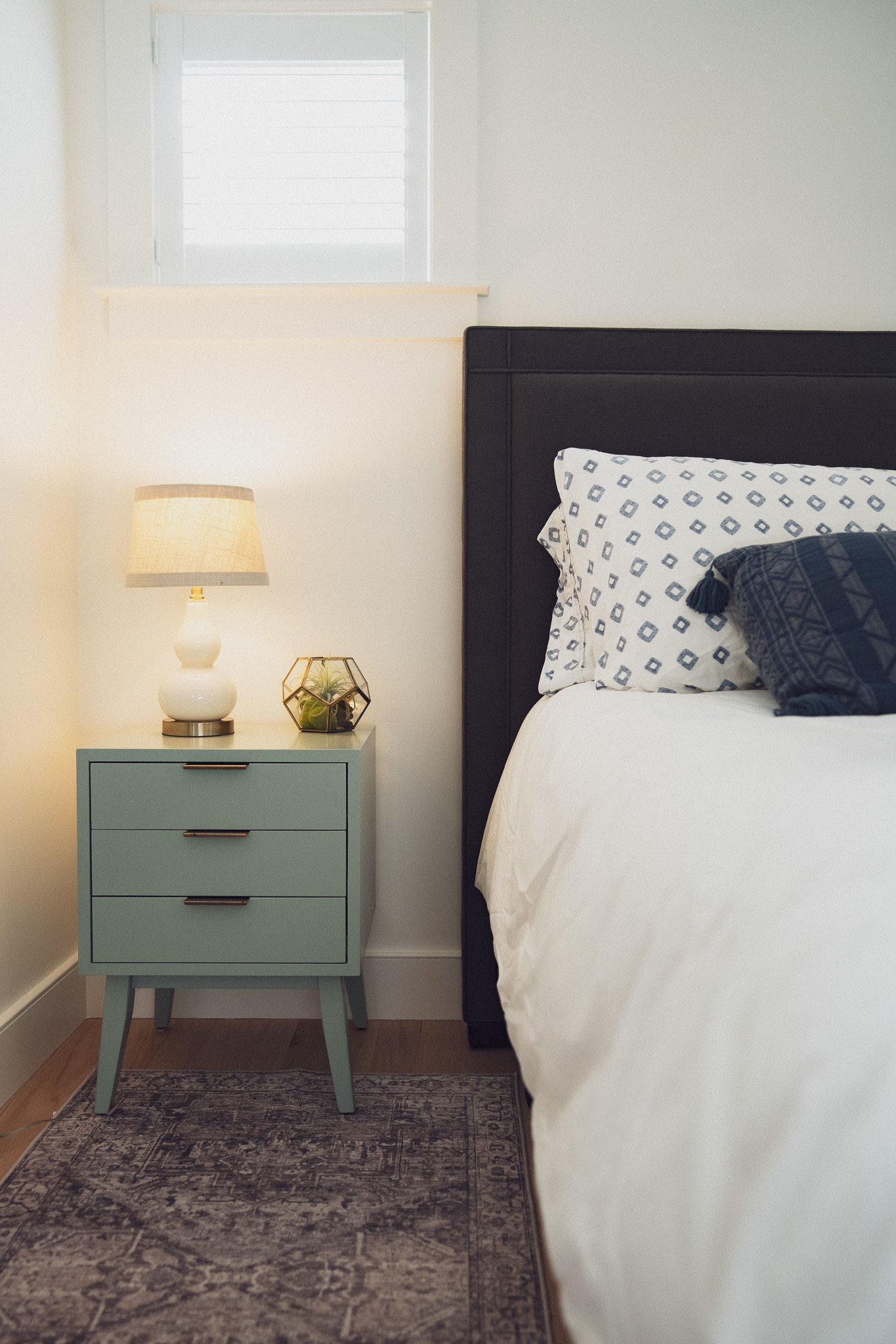 A cozy bedroom corner featuring a black headboard, white bed linens, blue patterned throw pillows, a light blue nightstand with three drawers, a white table lamp, and a decorative glass terrarium with plants on the nightstand. A small window with whi