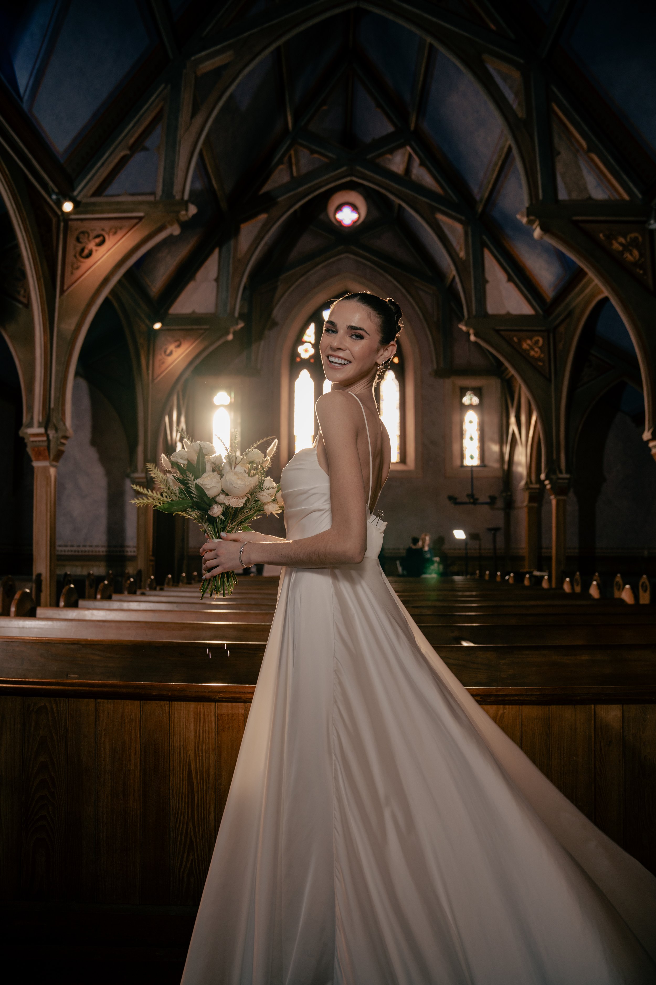 A smiling bride in a white wedding gown holding a bouquet of flowers inside a church with stained glass windows and gothic architecture.