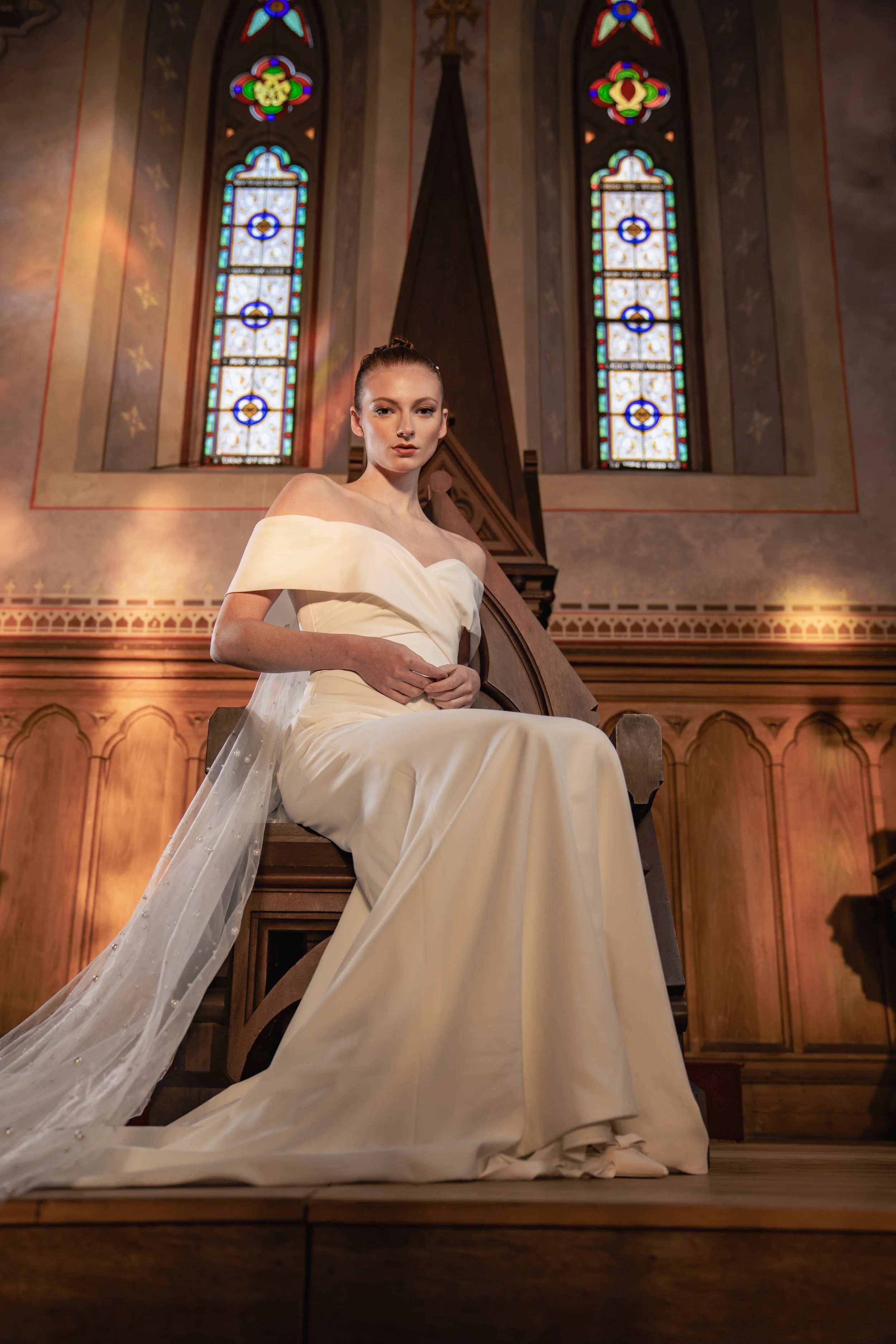 A woman in a white wedding dress sitting on a wooden chair inside a church with stained glass windows.