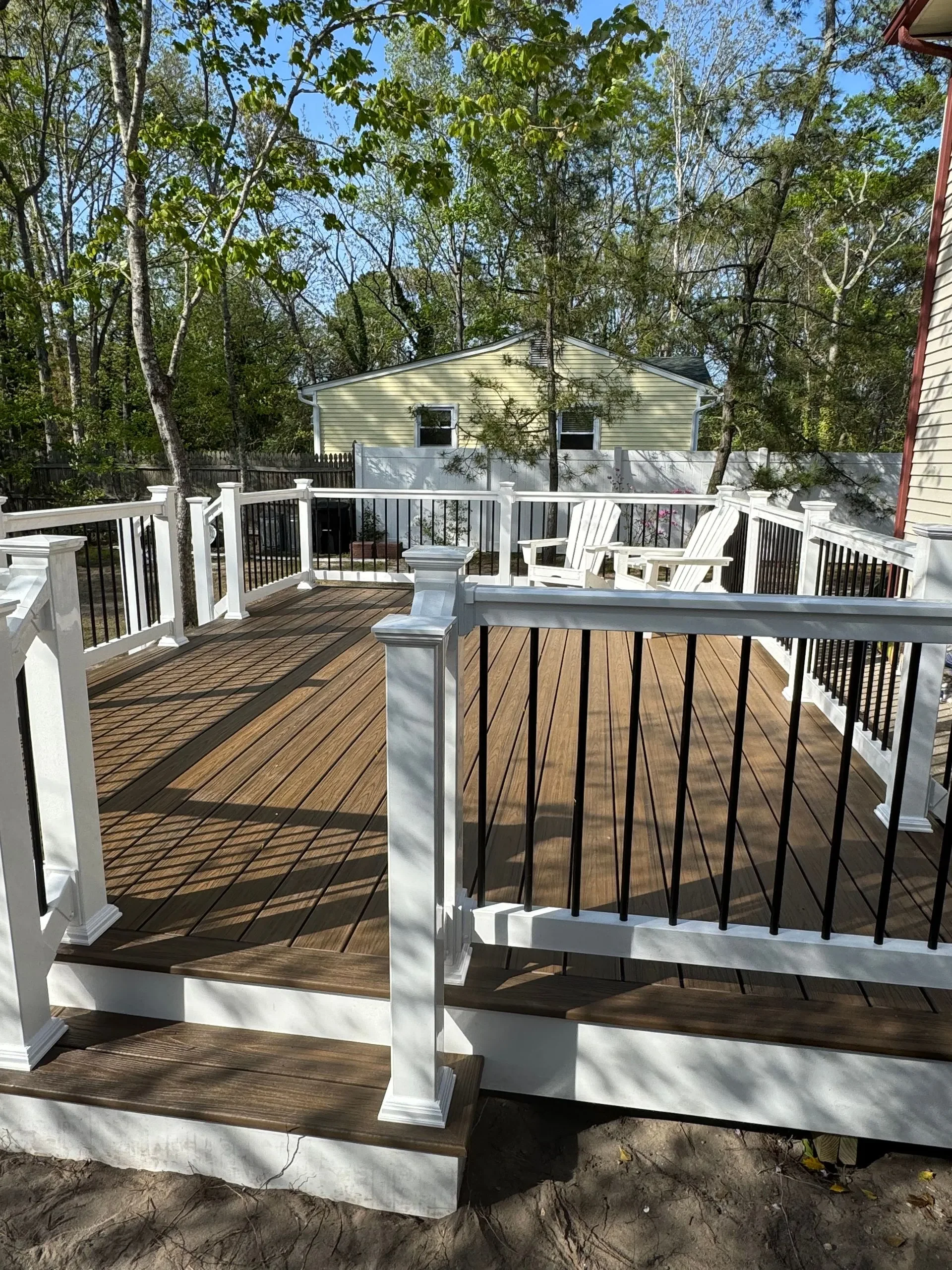 View of a newly built wooden backyard deck with white and black railing, two white Adirondack chairs, and trees in the background on a sunny day.