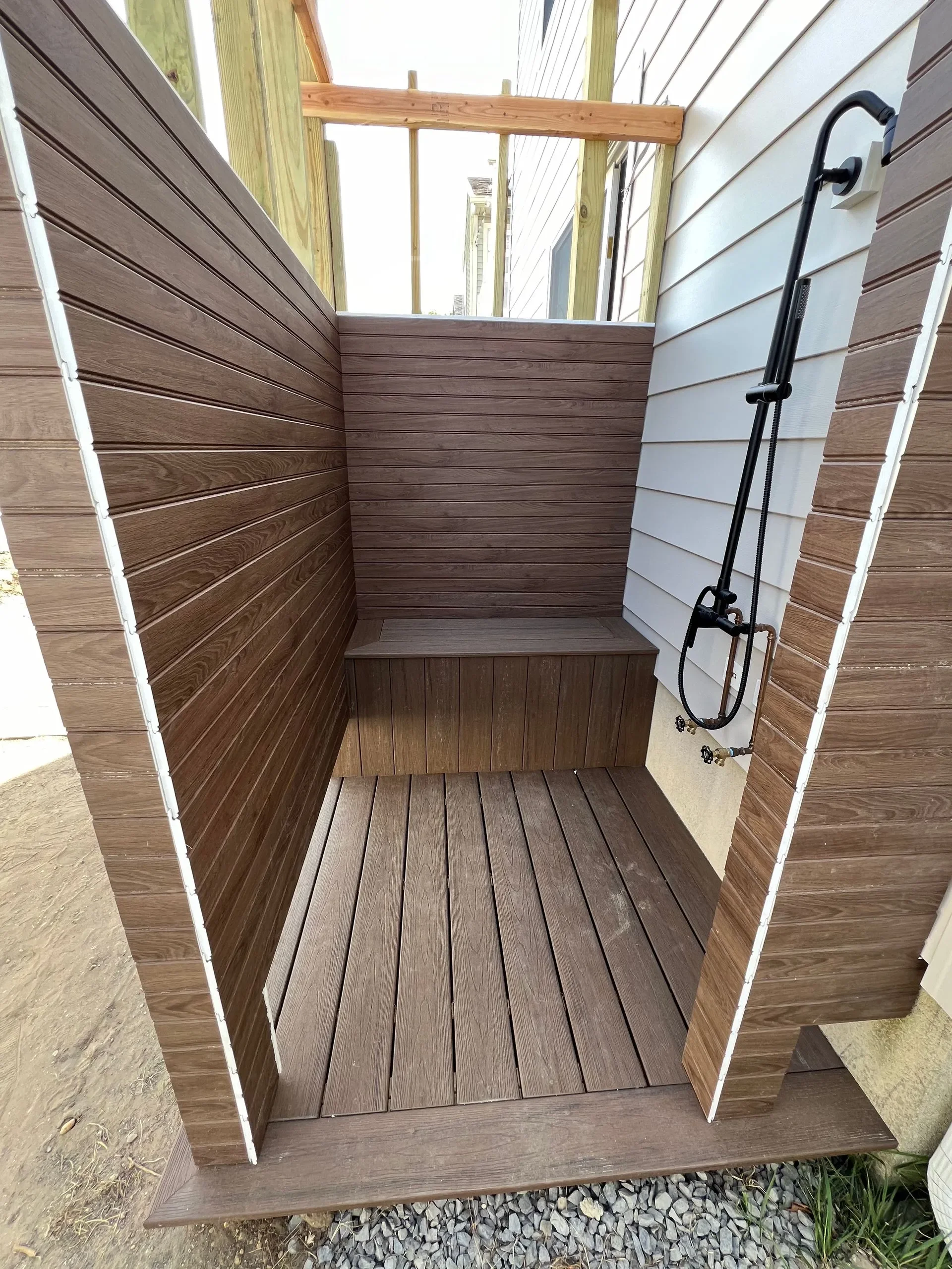 Outdoor shower with wood paneling and a black showerhead, bench, and floor with gravel at the base.