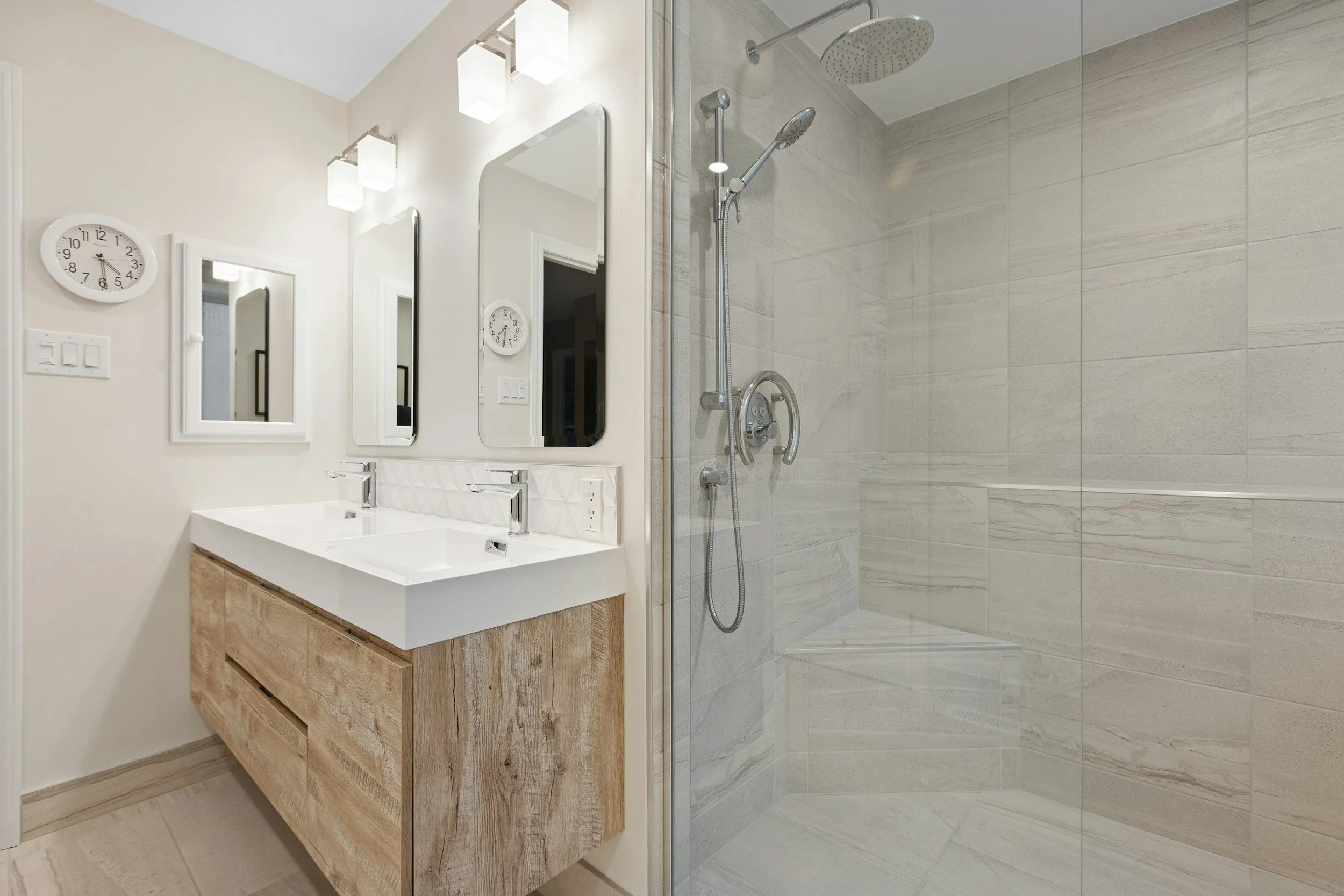 Modern bathroom with double sink vanity, large mirrors, wall-mounted lights, wall clock, and a walk-in shower with tiled walls and a rain showerhead.