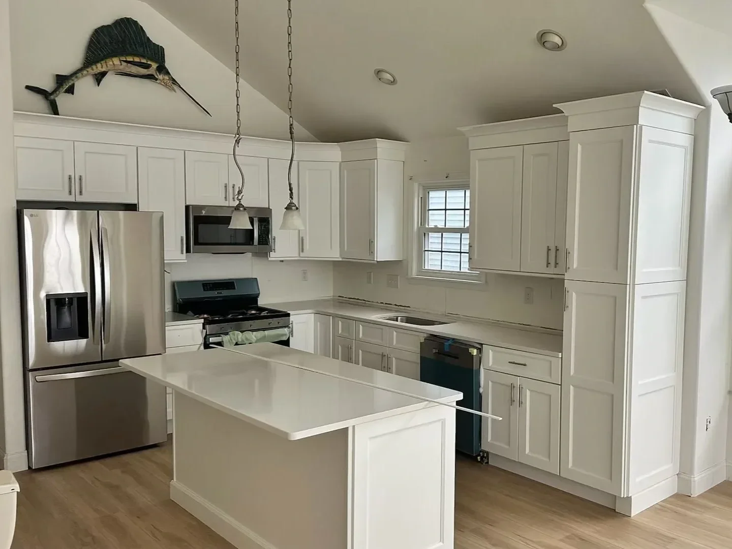 Empty kitchen with white cabinets, stainless steel refrigerator, microwave, stovetop, kitchen island, and window.