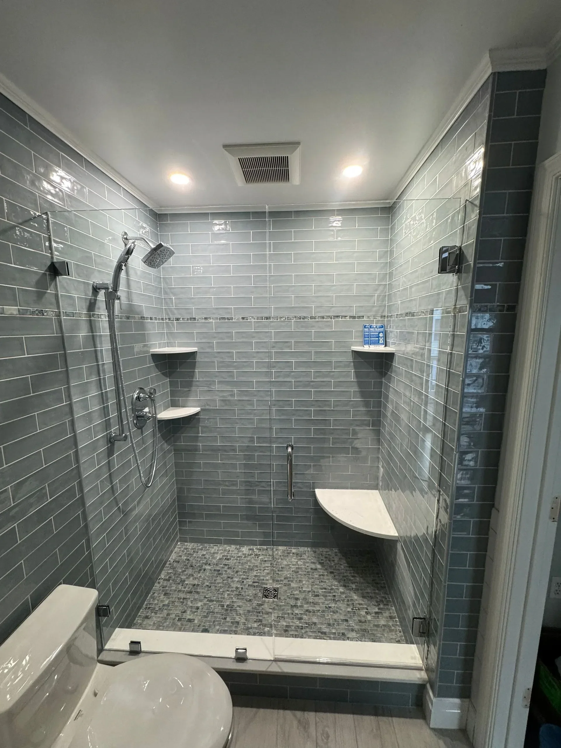 Modern walk-in shower with gray subway tile walls, a glass door, and built-in corner shelves. The shower has a handheld showerhead and two upper recessed lights, with a small bench and a grab bar for accessibility.
