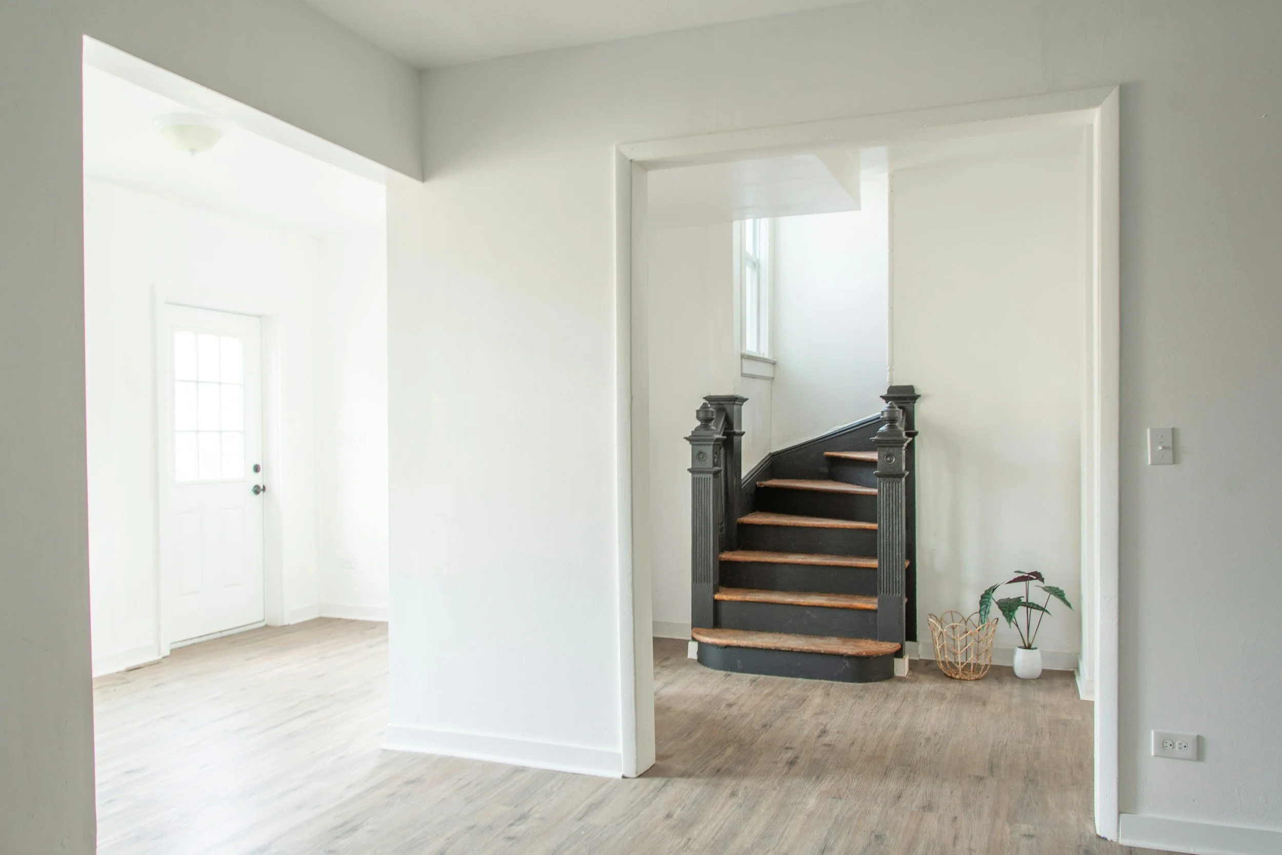 Interior of a house with white walls, a staircase with dark wood and black accents, a potted plant, and a basket on a wooden floor.