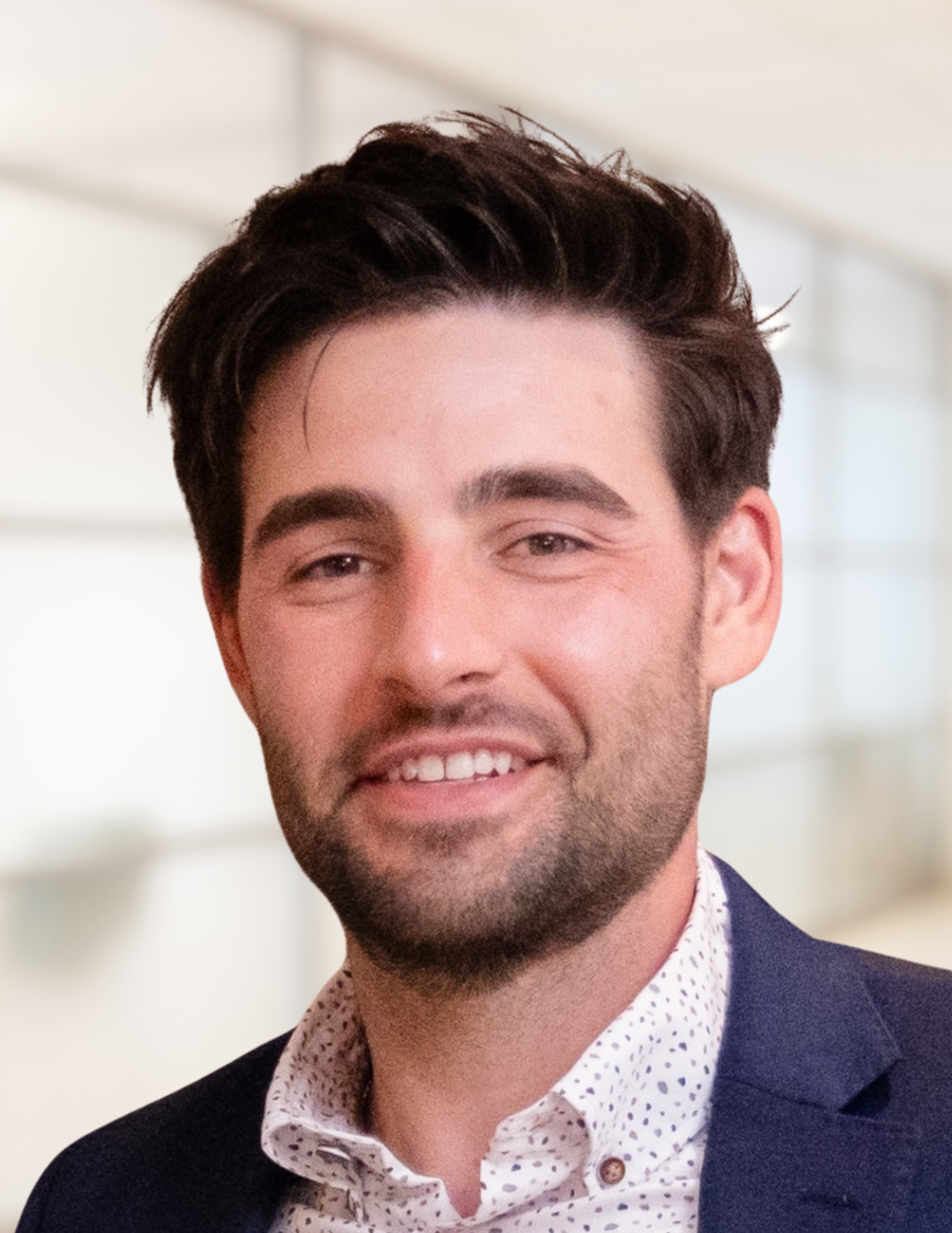 A smiling young man with dark hair, wearing a patterned white shirt and a navy blazer, indoors with a blurred background.