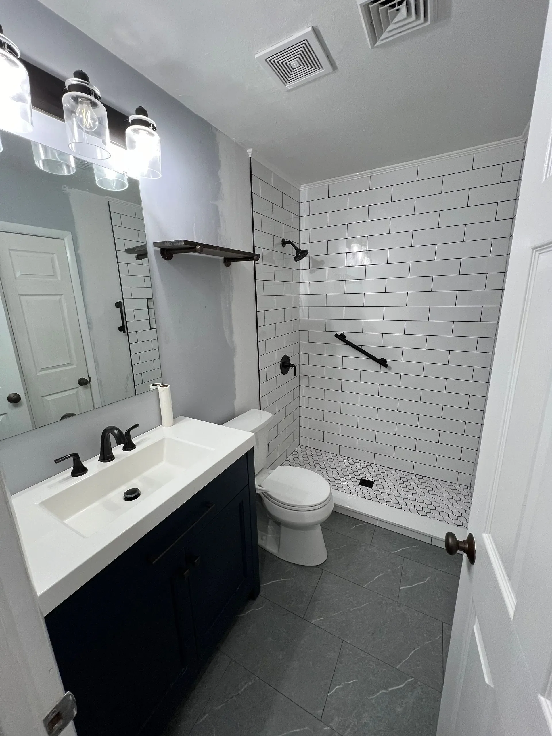 A modern bathroom with a walk-in shower featuring white subway tile walls, a black showerhead, and a black grab bar. There is a white toilet next to the shower and a black vanity with a white sink and black fixtures. A large mirror and a small black 