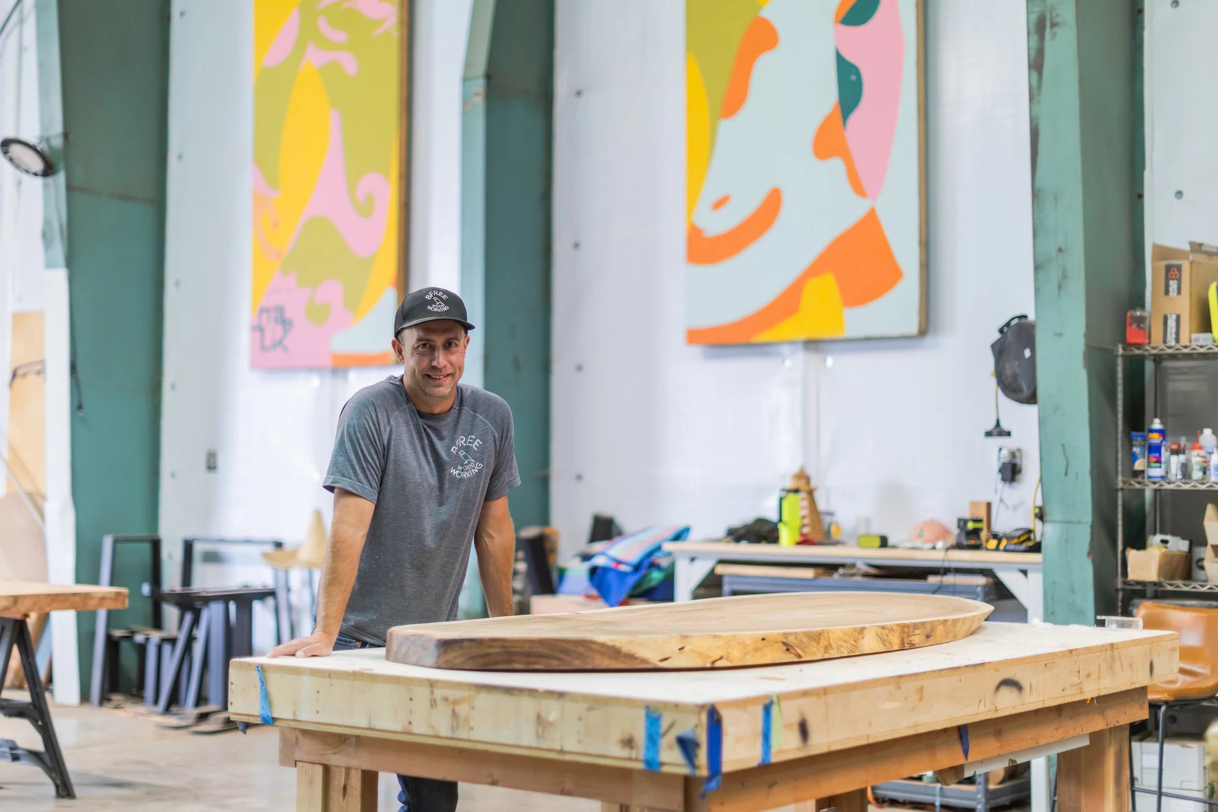 A man in a gray t-shirt and black cap leaning on a large piece of wood in a workshop with colorful abstract art on the walls and various tools and supplies around.