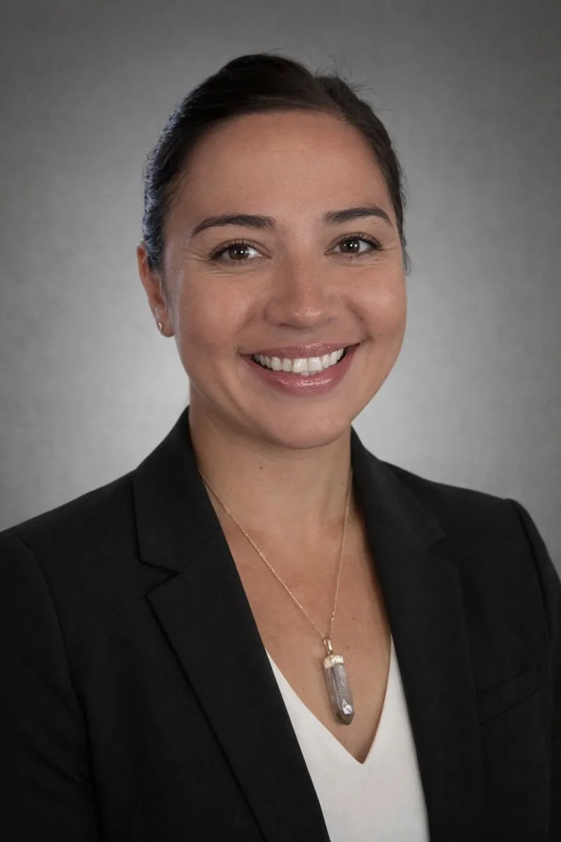 A woman with dark brown hair tied back, wearing a black blazer, white top, and a necklace with a large pendant, smiling at the camera against a gray background.