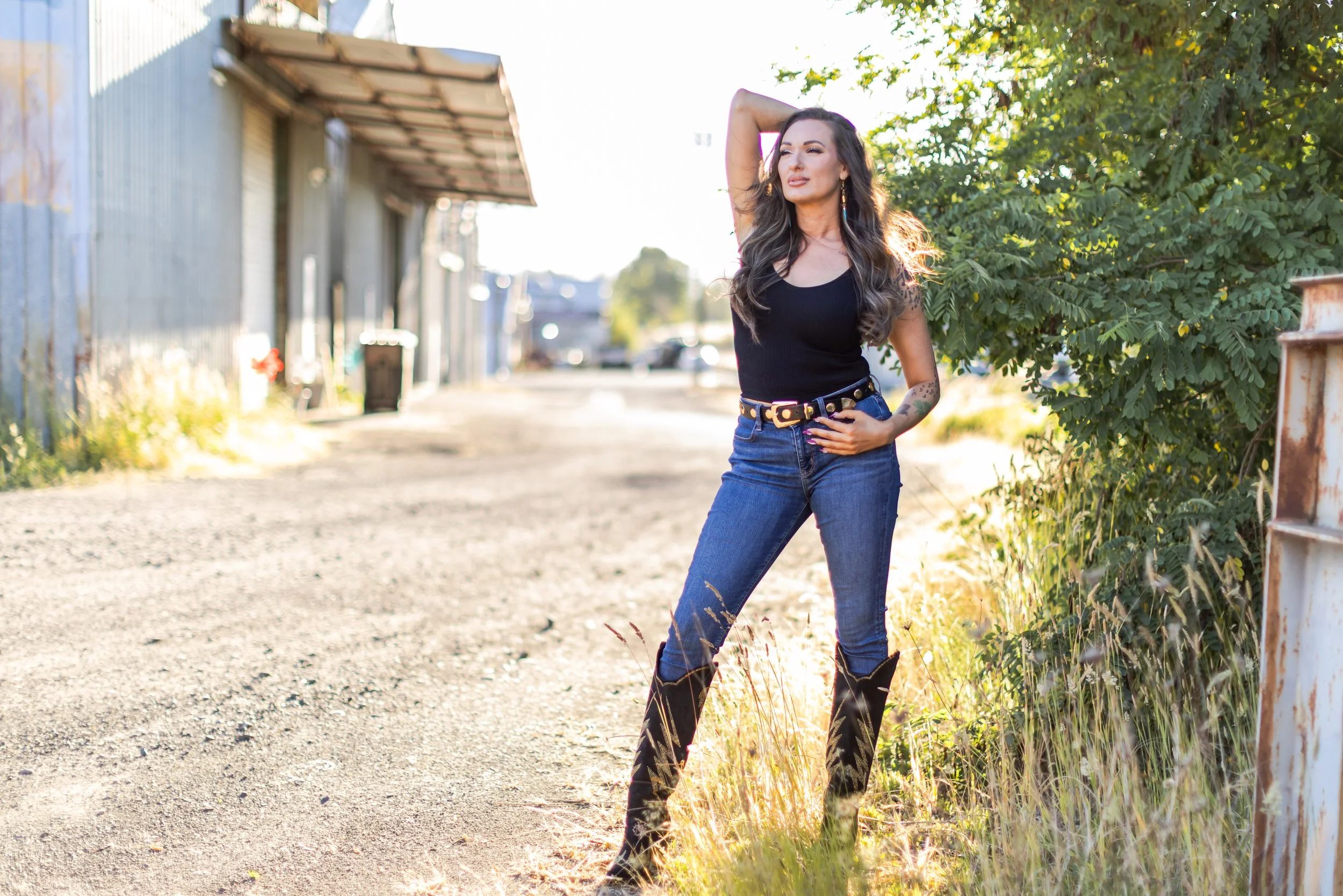 A woman with long wavy hair wearing a black sleeveless top, blue jeans, and cowboy boots standing outdoors next to a tree and rusty metal fence on a sunny day.