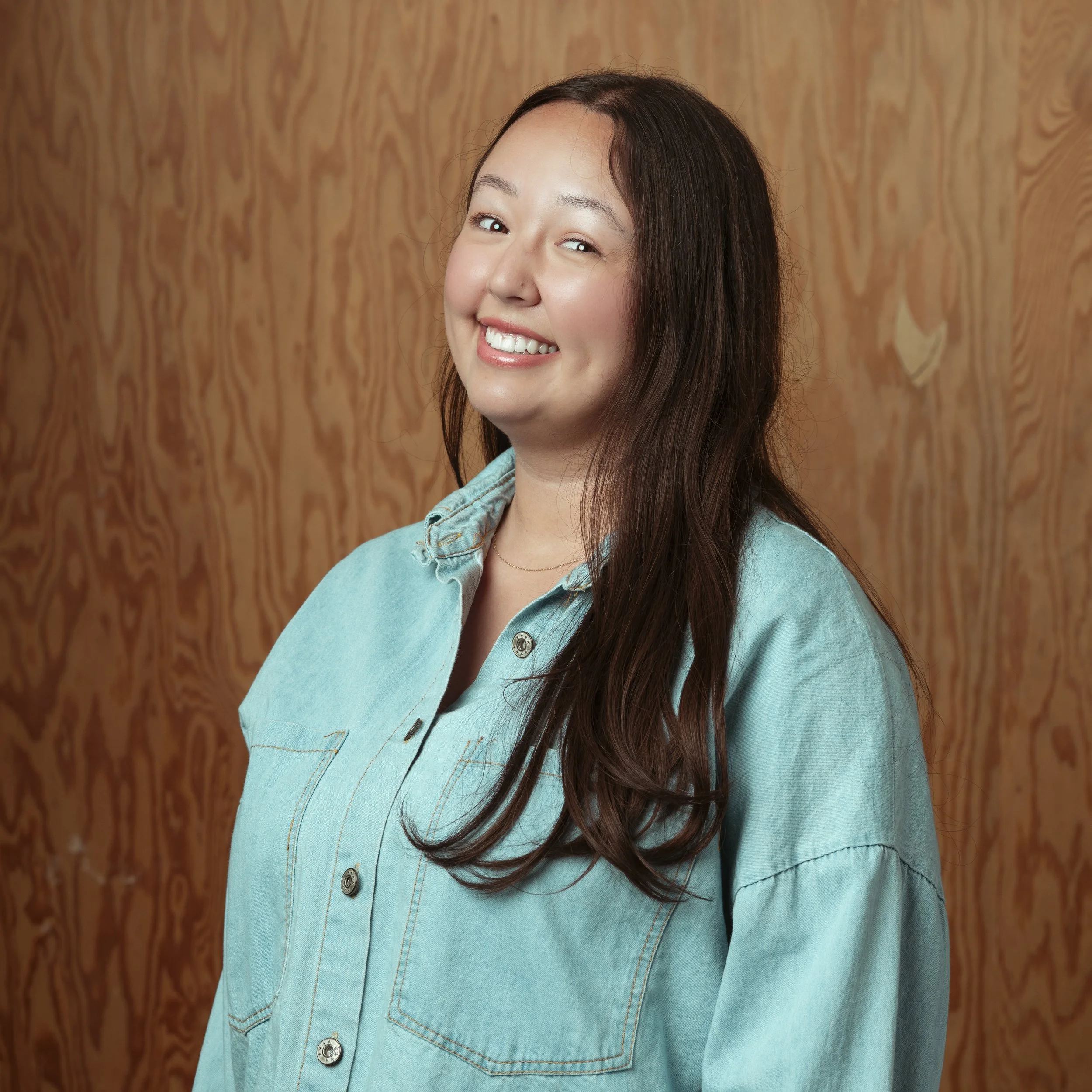 A woman with long brown hair, smiling with a slight head tilt, wearing a teal button-up shirt, standing in front of a wood-paneled wall.