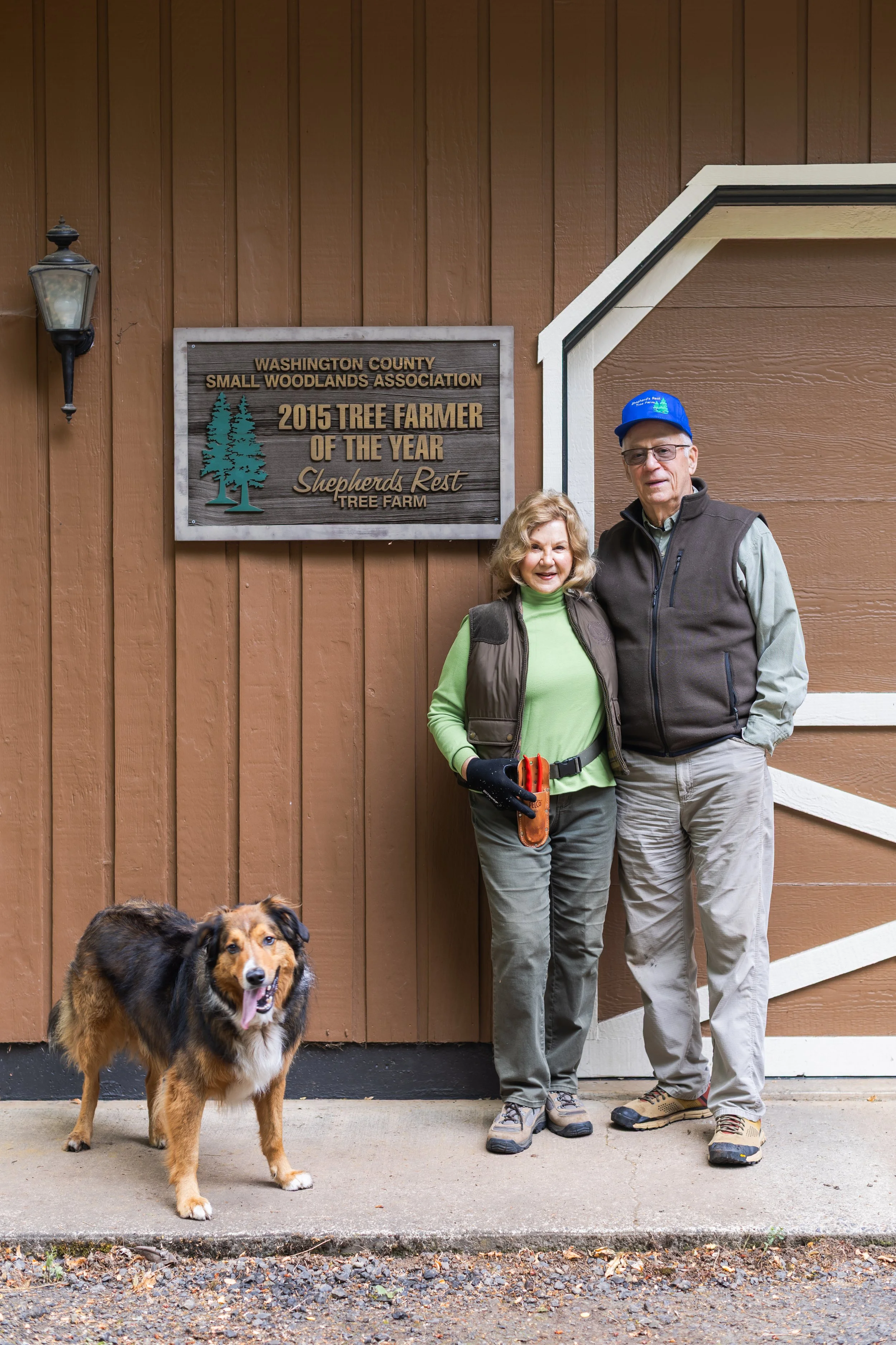A man and a woman are standing in front of a brown wooden building with a sign that reads "Washington County Small Woodlands Association, 2015 Tree Farmer of the Year, Shepherds Rest Tree Farm." Both are wearing outdoor clothing and smiling at the ca