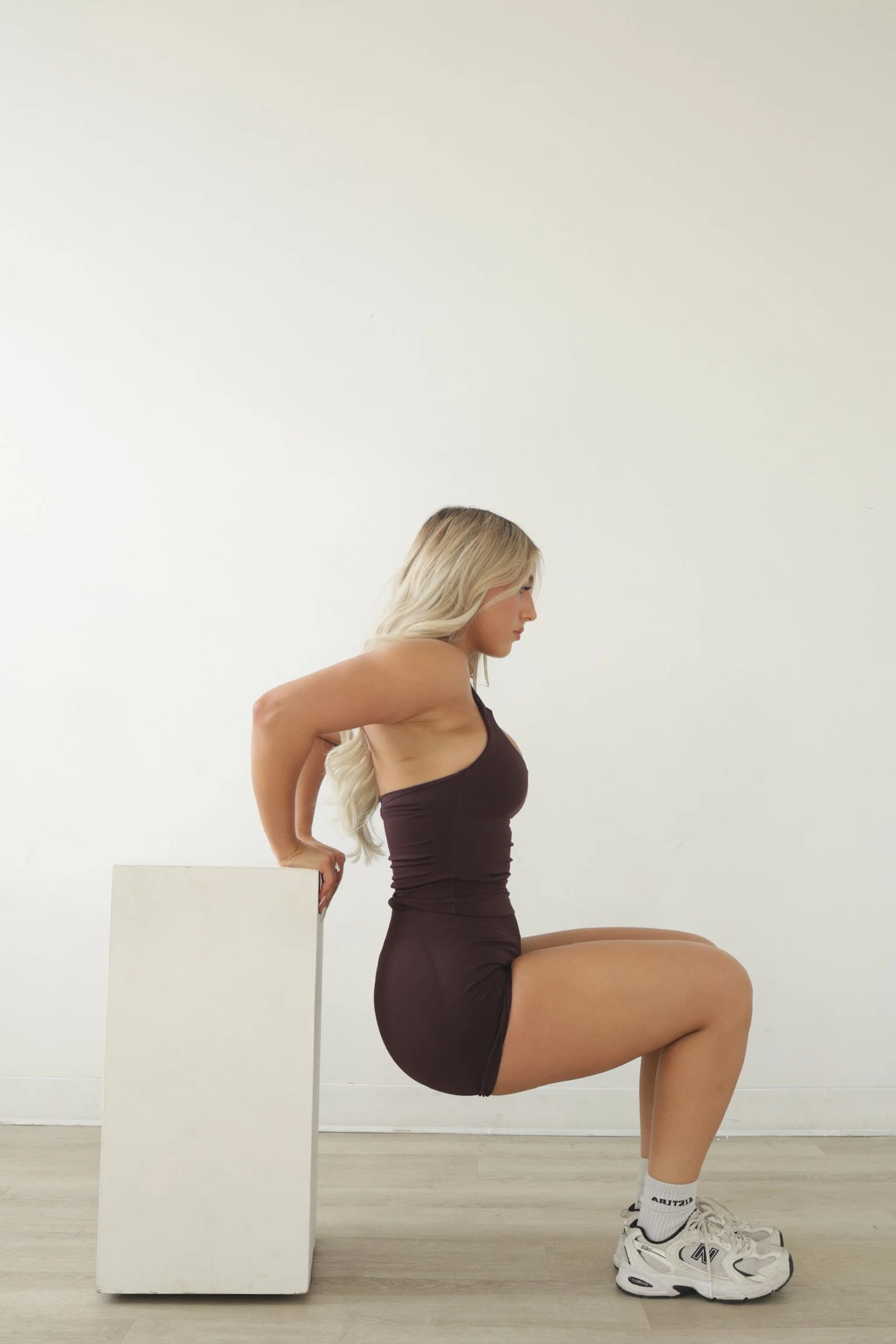 A woman with long blonde hair performing a tricep dip exercise on a white rectangular block in a minimal indoor setting with a white wall and wooden floor.