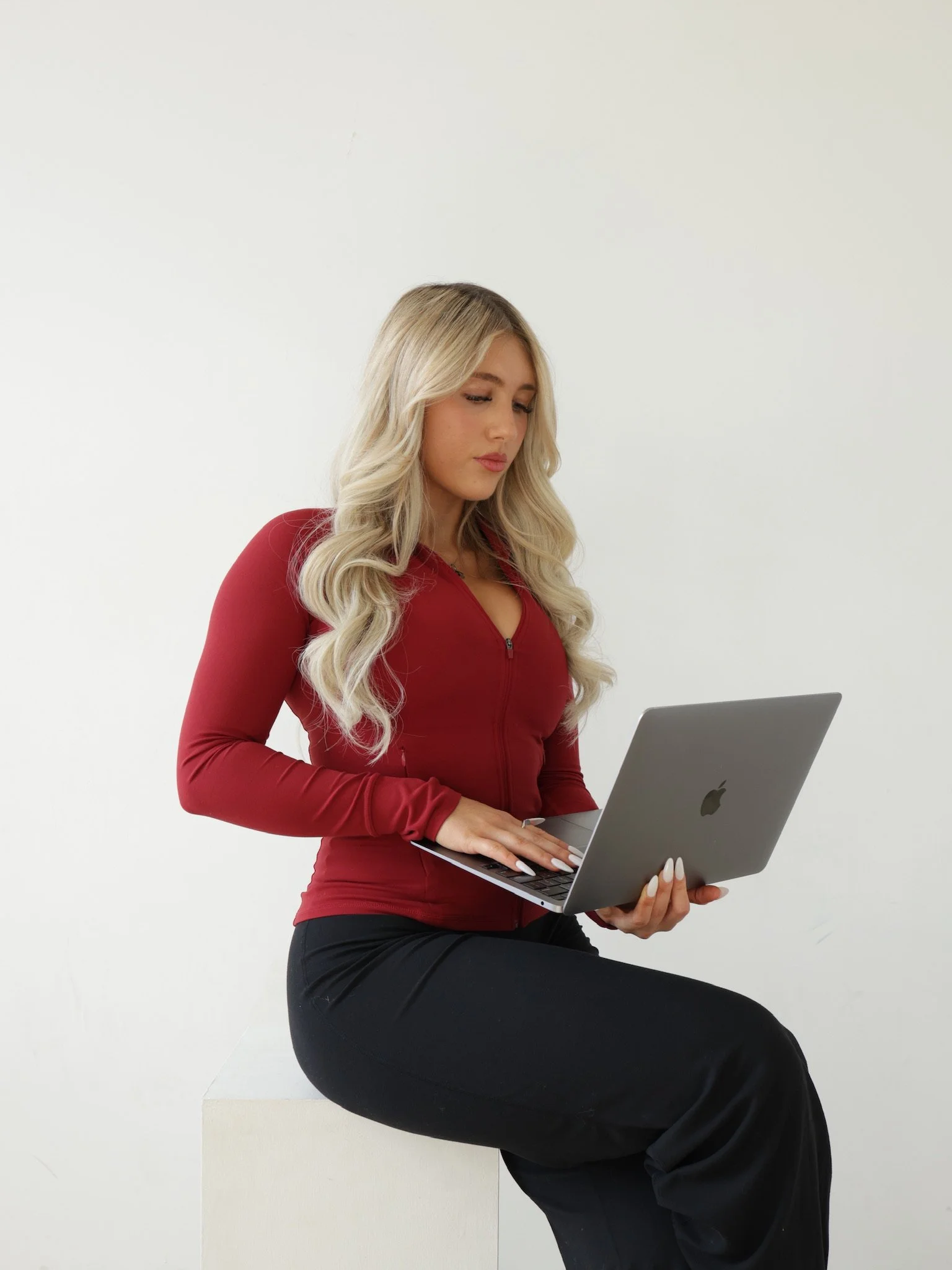 A woman with long blond hair, wearing a red zip-up top and black pants, sitting on a white block while holding and using a silver MacBook laptop against a plain white background.