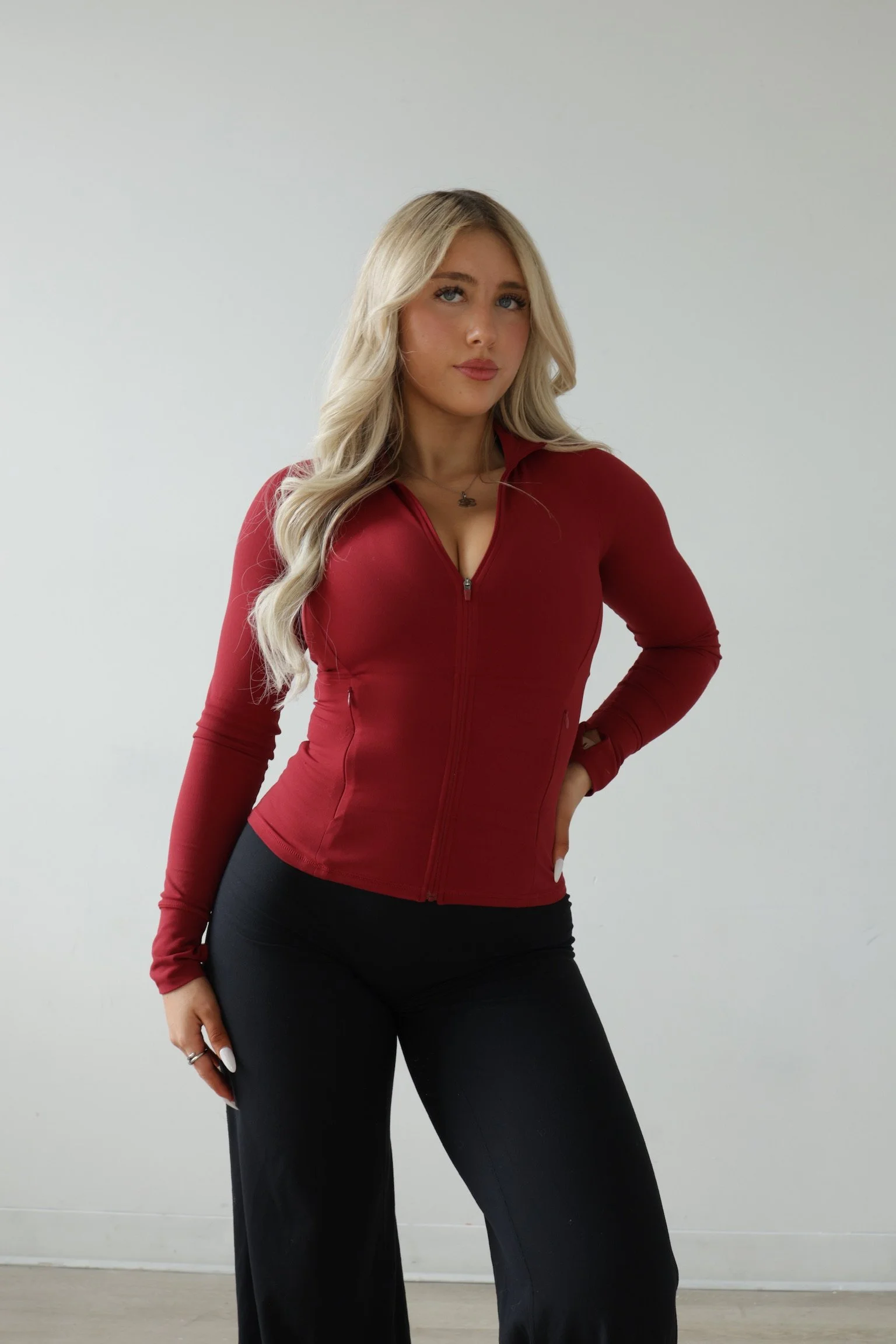 A woman with long blonde hair wearing a red zip-up jacket and black pants, posing with one hand on her hip against a plain white wall.