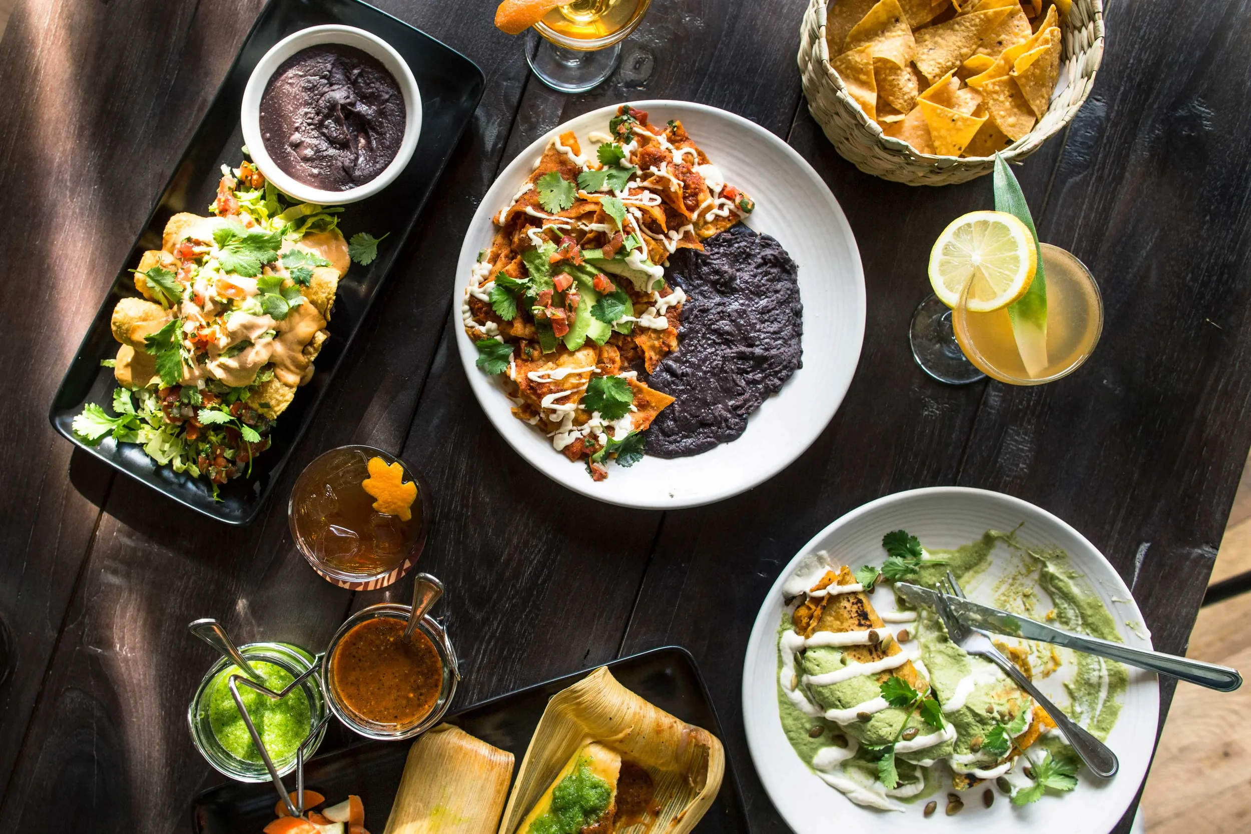 Assorted Mexican dishes on a dark wooden table, including tacos with cilantro and sauce, black beans with rice, guacamole, tortilla chips with salsa, and drinks with lemon and lime.