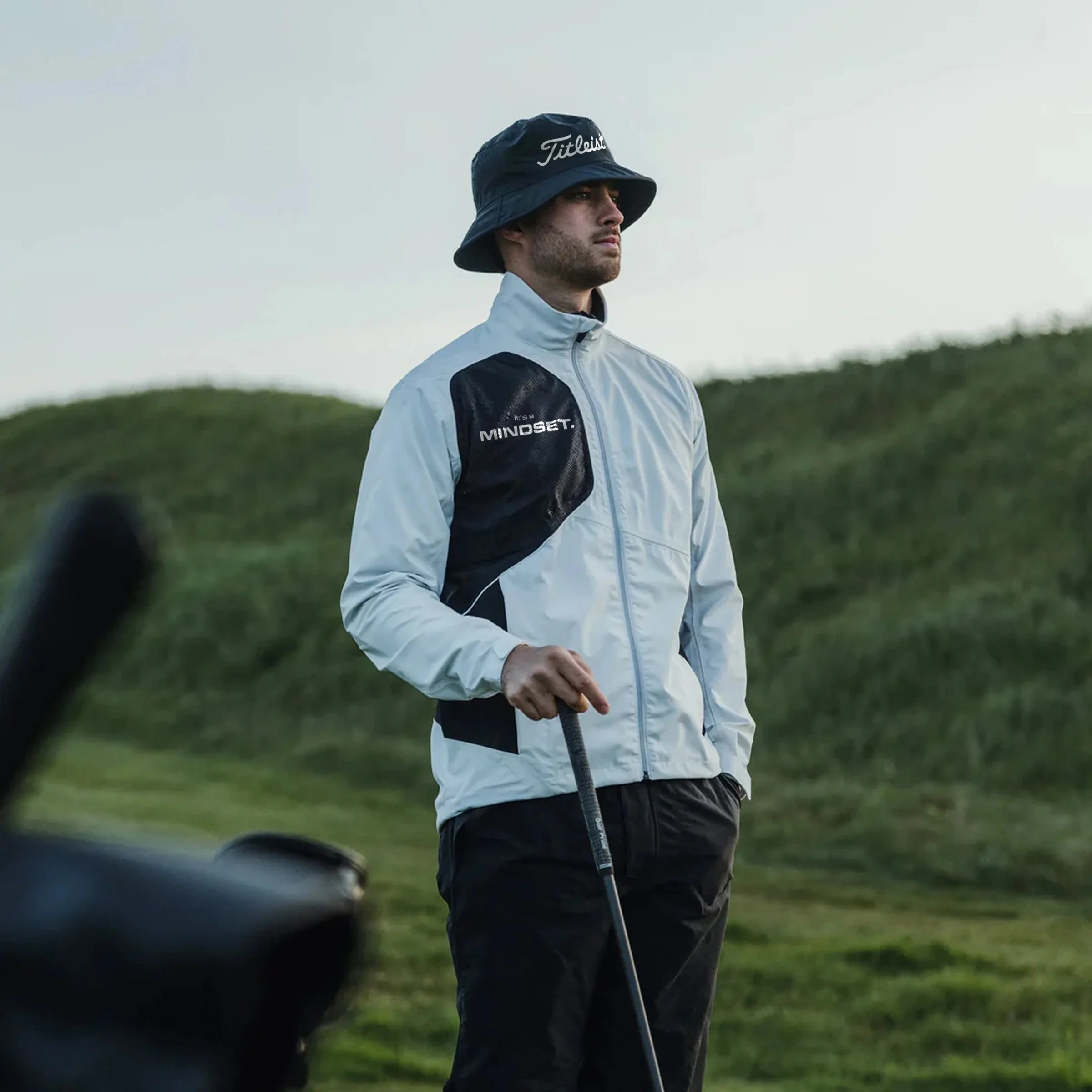 A man wearing a black bucket hat, white jacket, and black pants holding a golf club on a golf course, with green hills in the background.