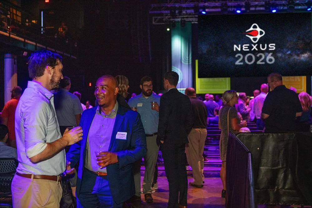People at a professional networking event with a large screen displaying 'Nexus 2026' in the background.