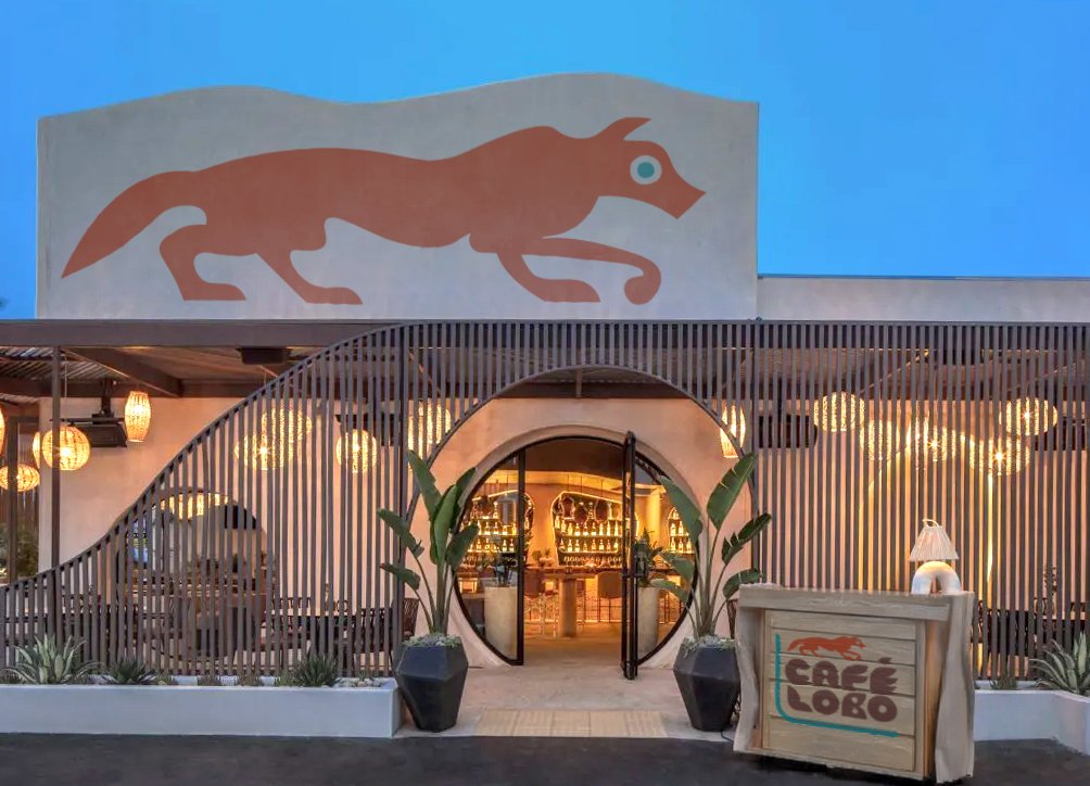 Exterior of a cafe named 'Cafe Lobo' with a stylized wolf logo on a sign above the entrance, illuminated interior, potted plants, and warm lighting.