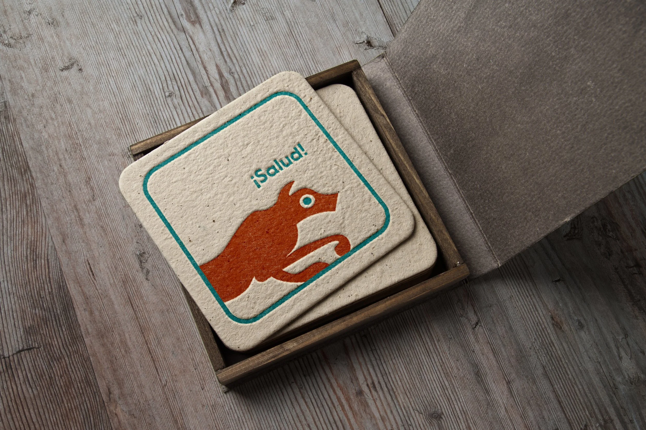 A set of coasters with a cartoon orange animal and the Spanish word '¡salud!', placed inside a wooden box on a wooden table.