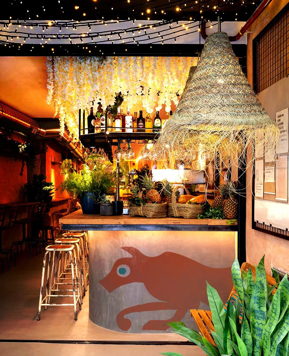 Decorated bar area with hanging string lights, a woven lampshade, and tropical plants, featuring a bar with a painted animal silhouette on the front and bottles on a high shelf, creating a cozy ambiance.