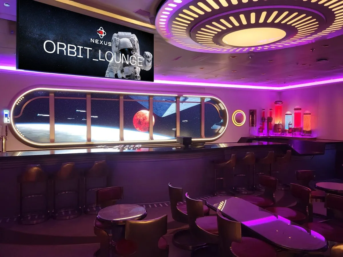 A futuristic space-themed lounge called 'Orbit Lounge'. It features a large window overlooking space with a view of Earth, a red moon, and spacecraft. The space is decorated with pink and purple lighting, round tables, chairs, and a counter with vari