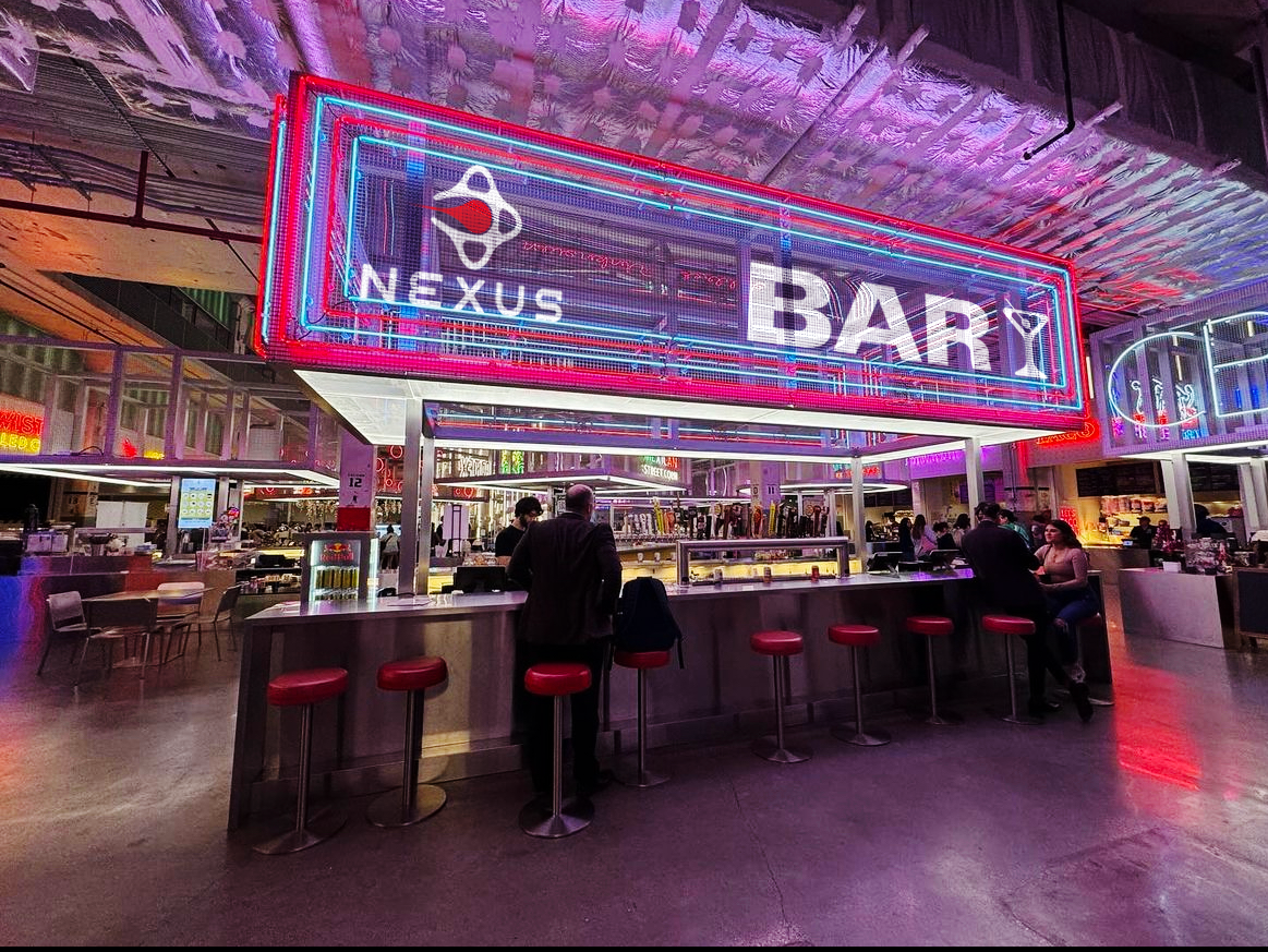 Neon sign for Nexus Bar inside a modern, colorful bar or food court with people sitting and standing around the counter.