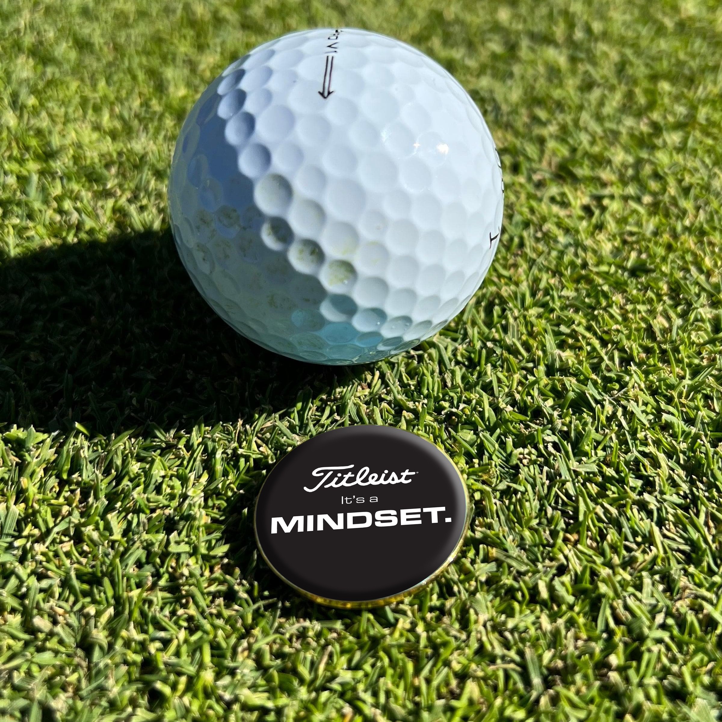 A golf ball on grass near a black button with the text 'Titleist It's a MINDSET.'