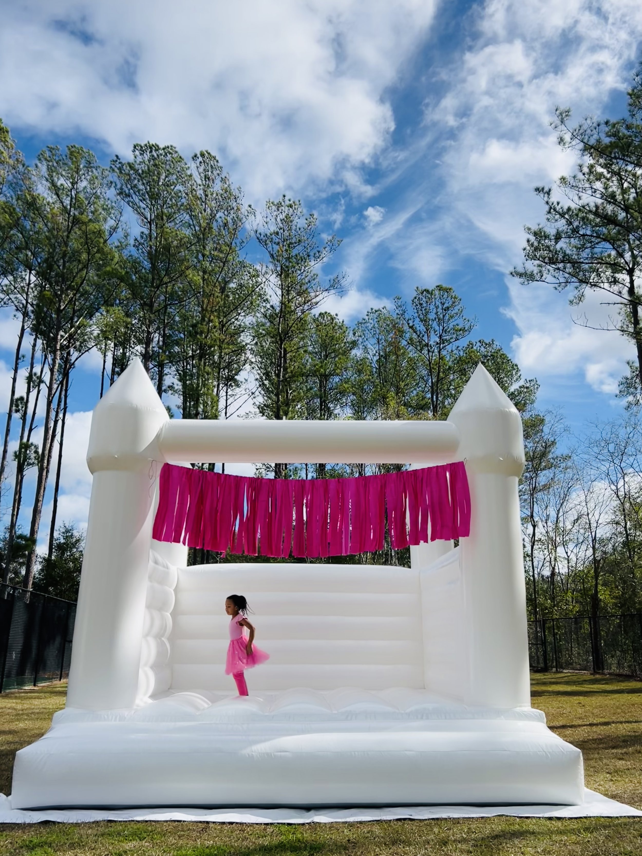 A young girl in a purple dress with a pink bow in her hair jumping inside a white inflatable bounce house that resembles a castle, under a cloudy sky.
