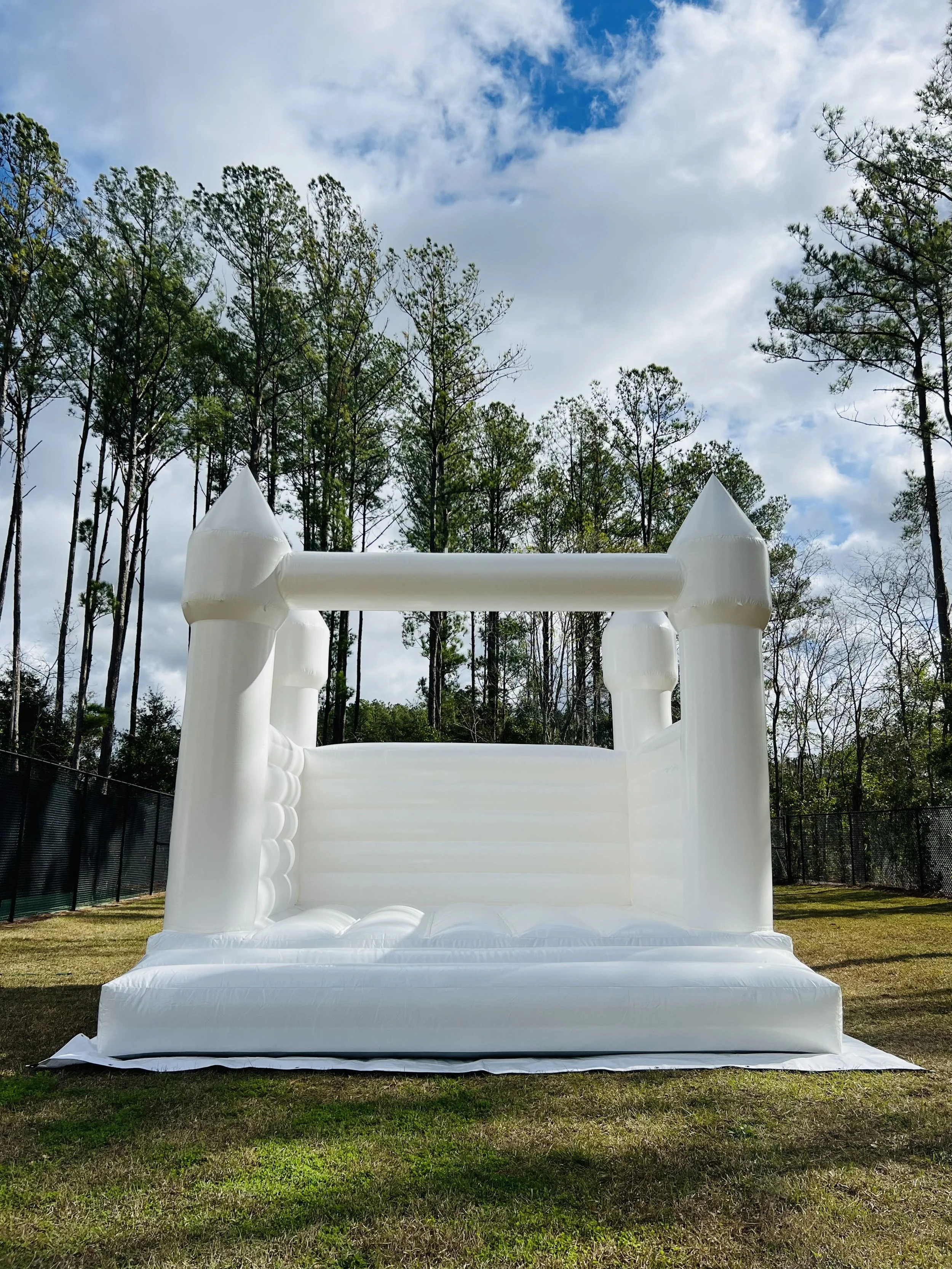 Inflatable white bounce house with castle-like towers on a grassy field surrounded by trees under a partly cloudy sky.