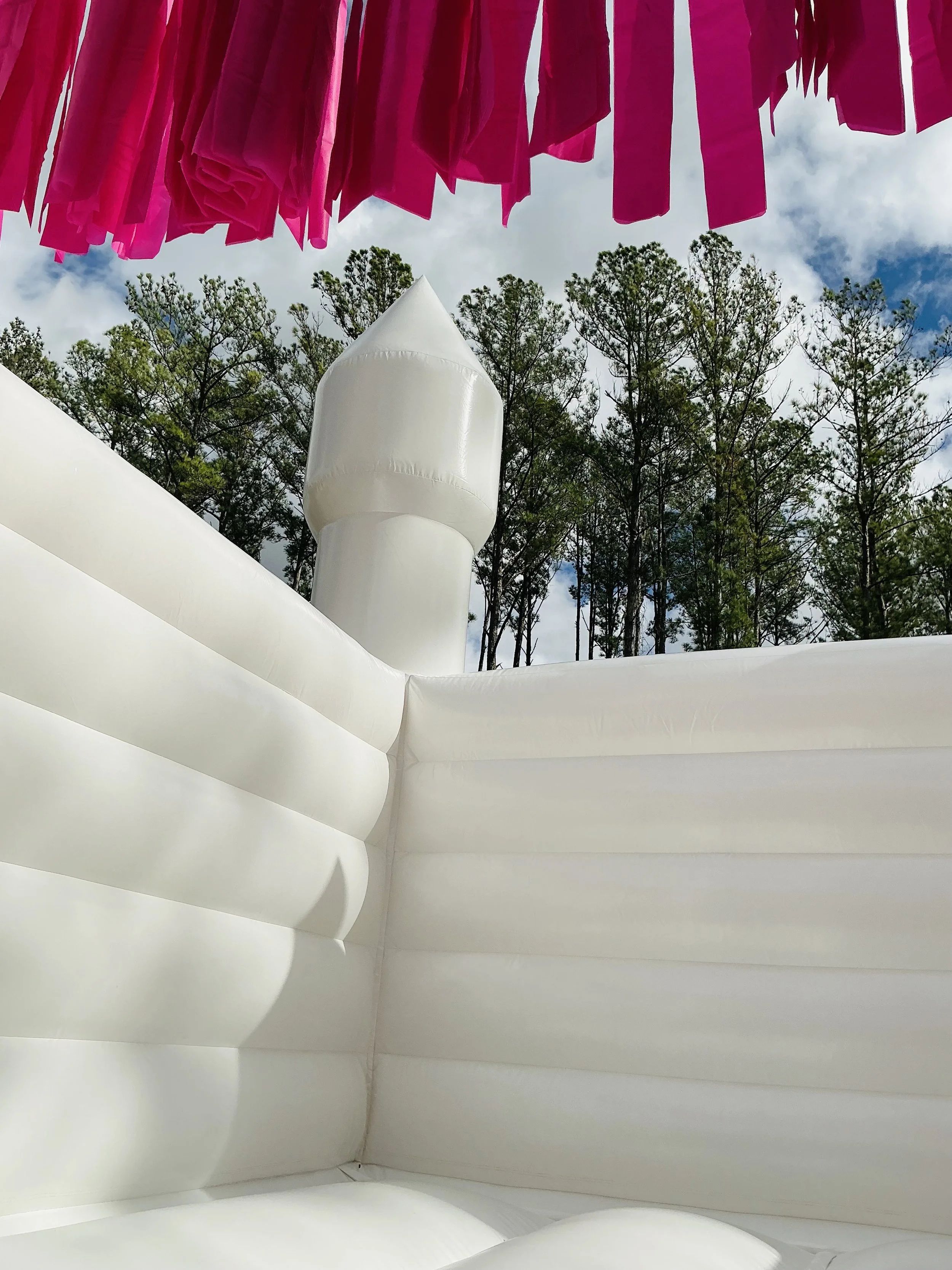 Inflatable white bounce house with a tower, seen outdoors with trees and a cloudy sky in the background, and pink fabric hanging overhead.