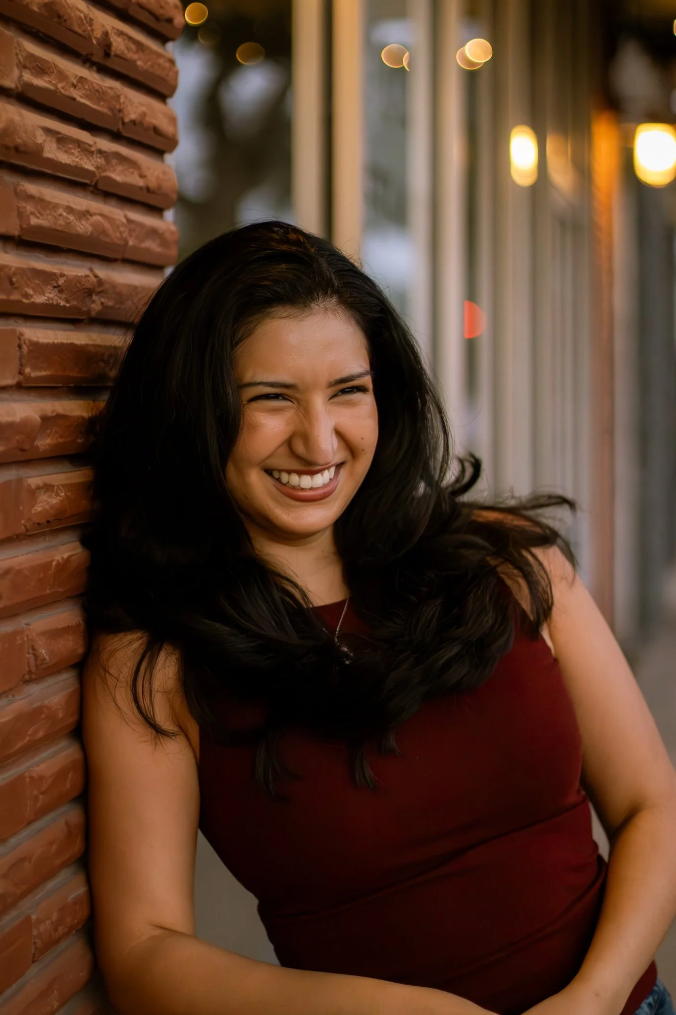 A young woman with long dark hair smiling and leaning against a brick wall outside, with blurred lights in the background.