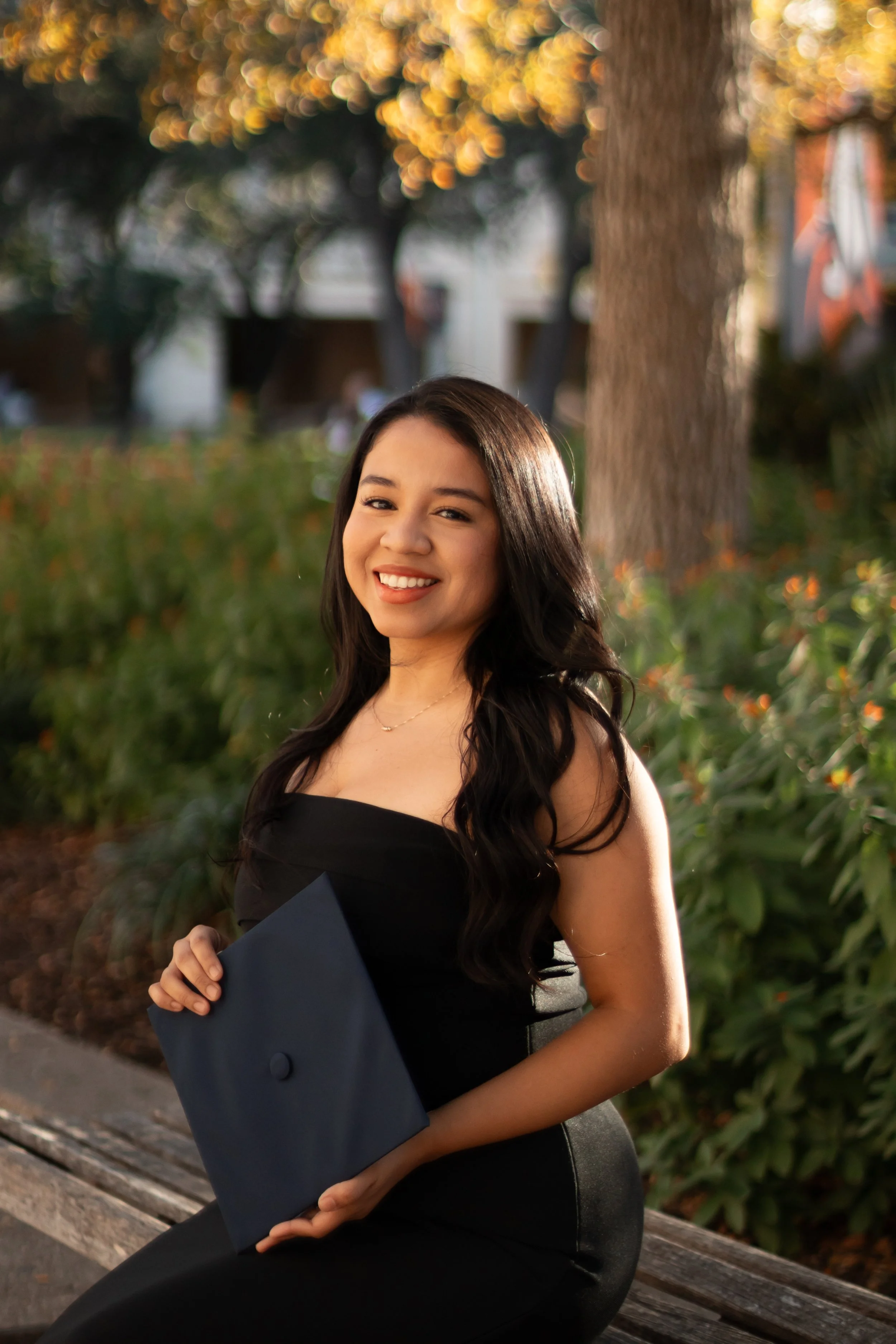A young woman in a black dress sitting on a park bench, holding a navy diploma cover, smiling happily with trees and blurred background in the evening light.